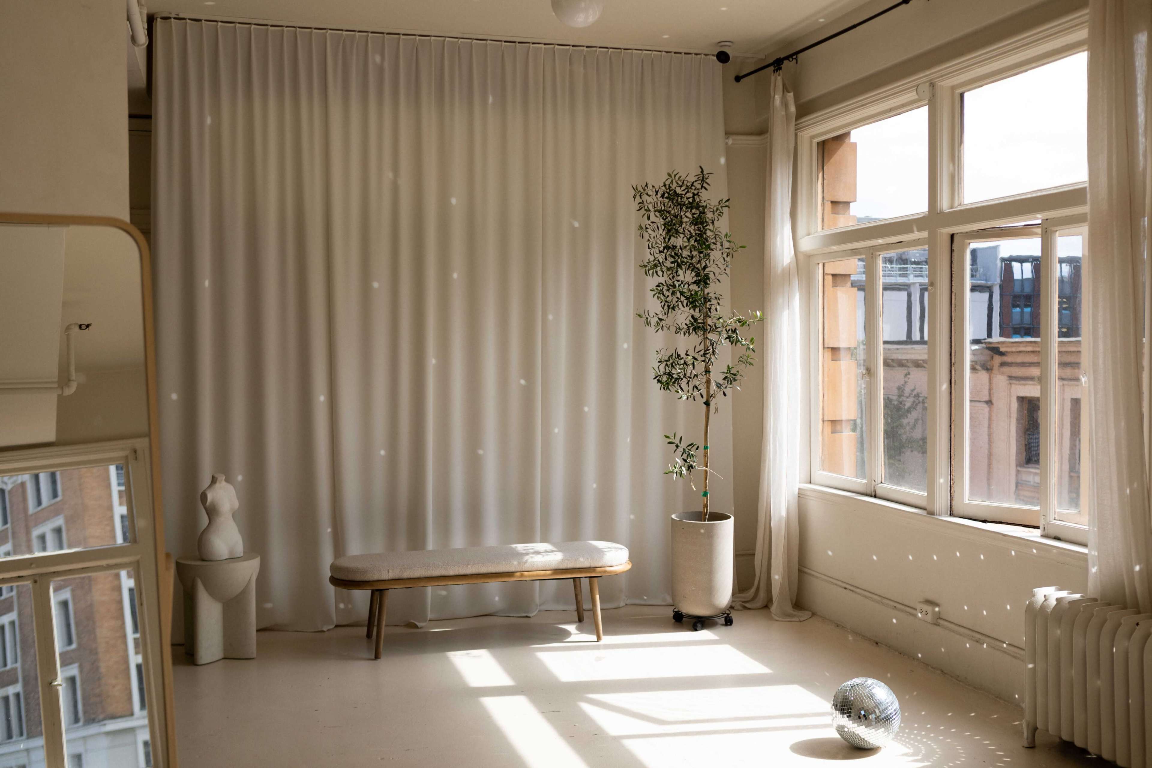 A sunlit room features a bench, a potted plant, floor-to-ceiling curtains, and a reflective sphere on the floor.