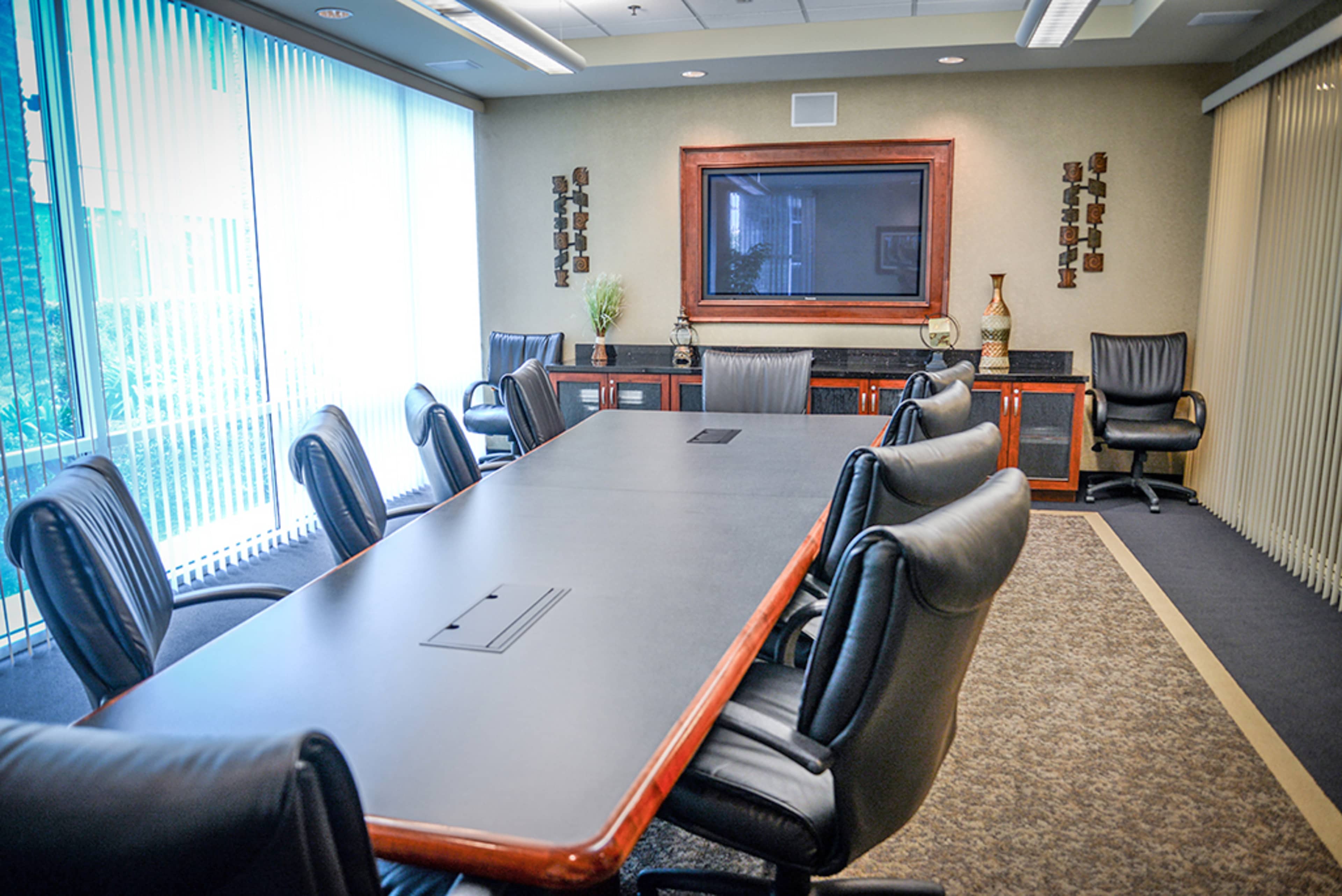A conference room is equipped with a long table surrounded by black leather chairs and features a large screen on the wall.