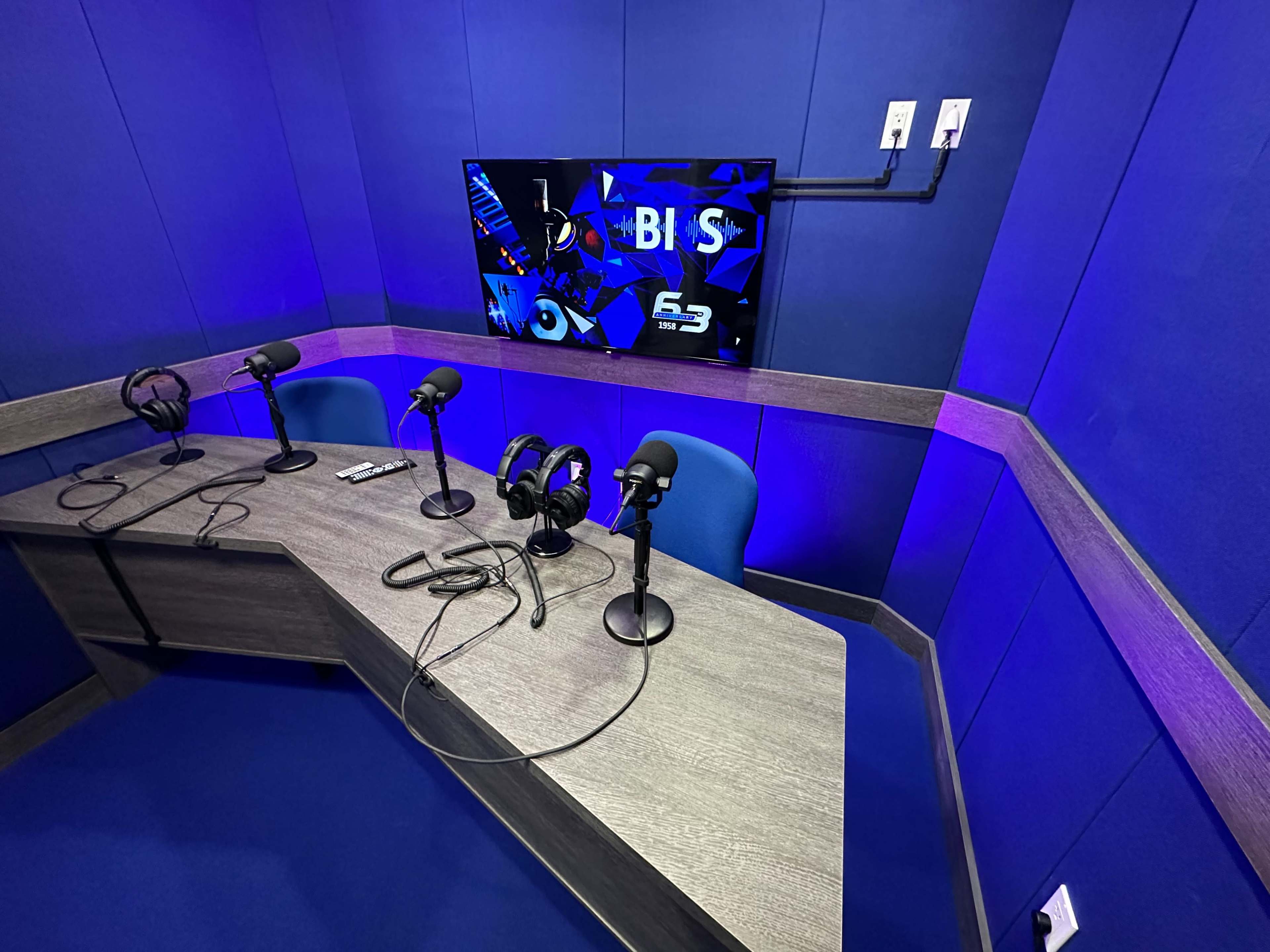 A recording studio with a blue backdrop, featuring a large monitor and three microphone setups on a wooden table.