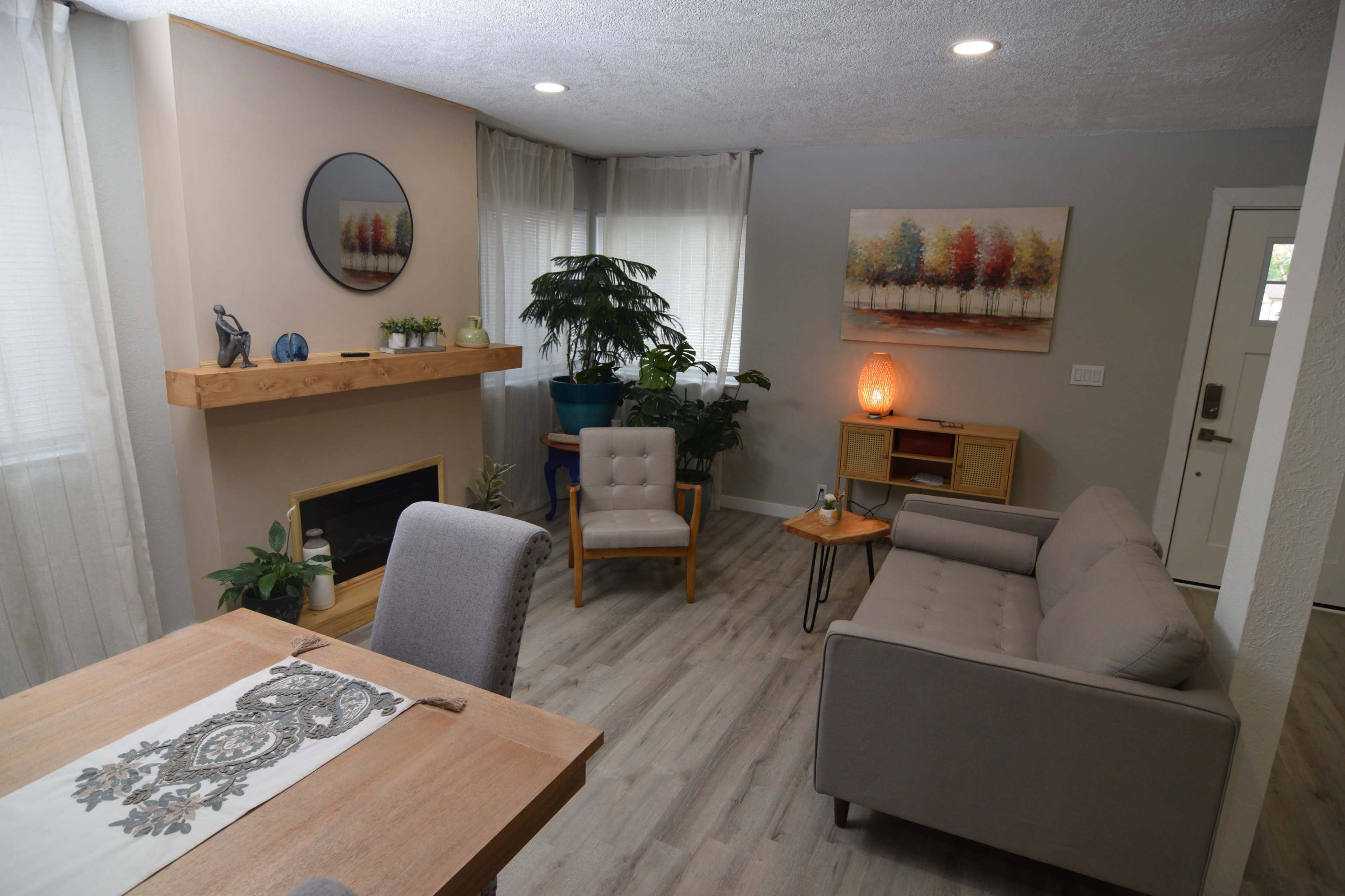 The living room features a sofa, two chairs, a wooden table, a fireplace, and a large window with sheer curtains, complemented by decorative plants and artwork on the walls.