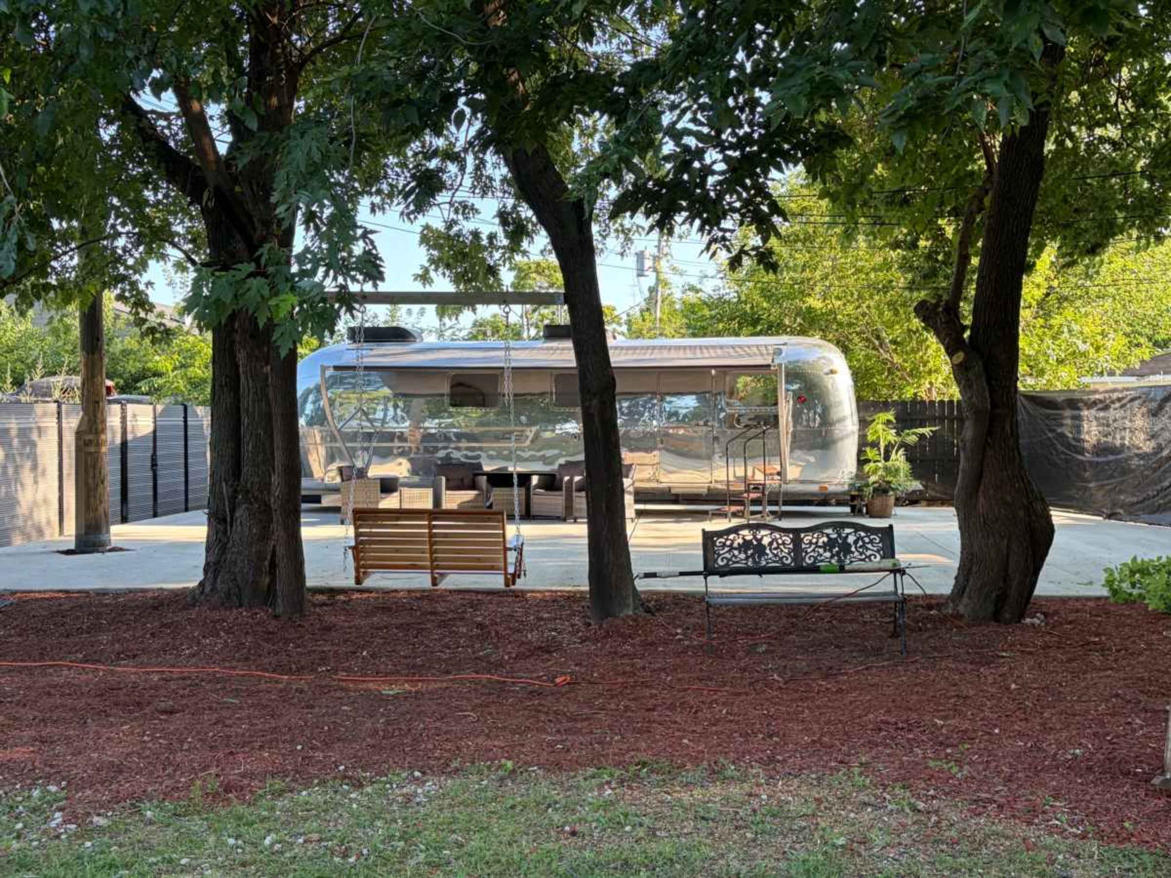 A polished Airstream trailer is parked on a paved area surrounded by trees, with two benches placed nearby.