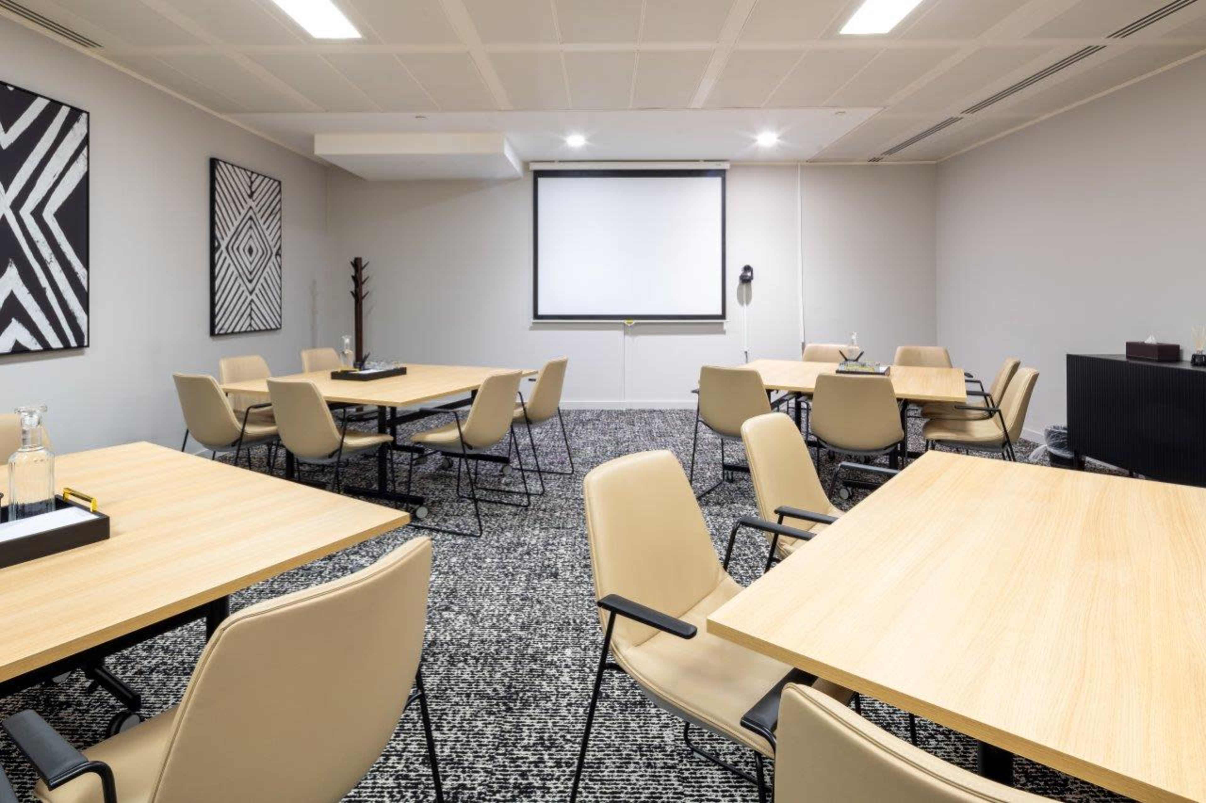 A modern meeting room with several wooden tables and beige chairs arranged around a central screen.