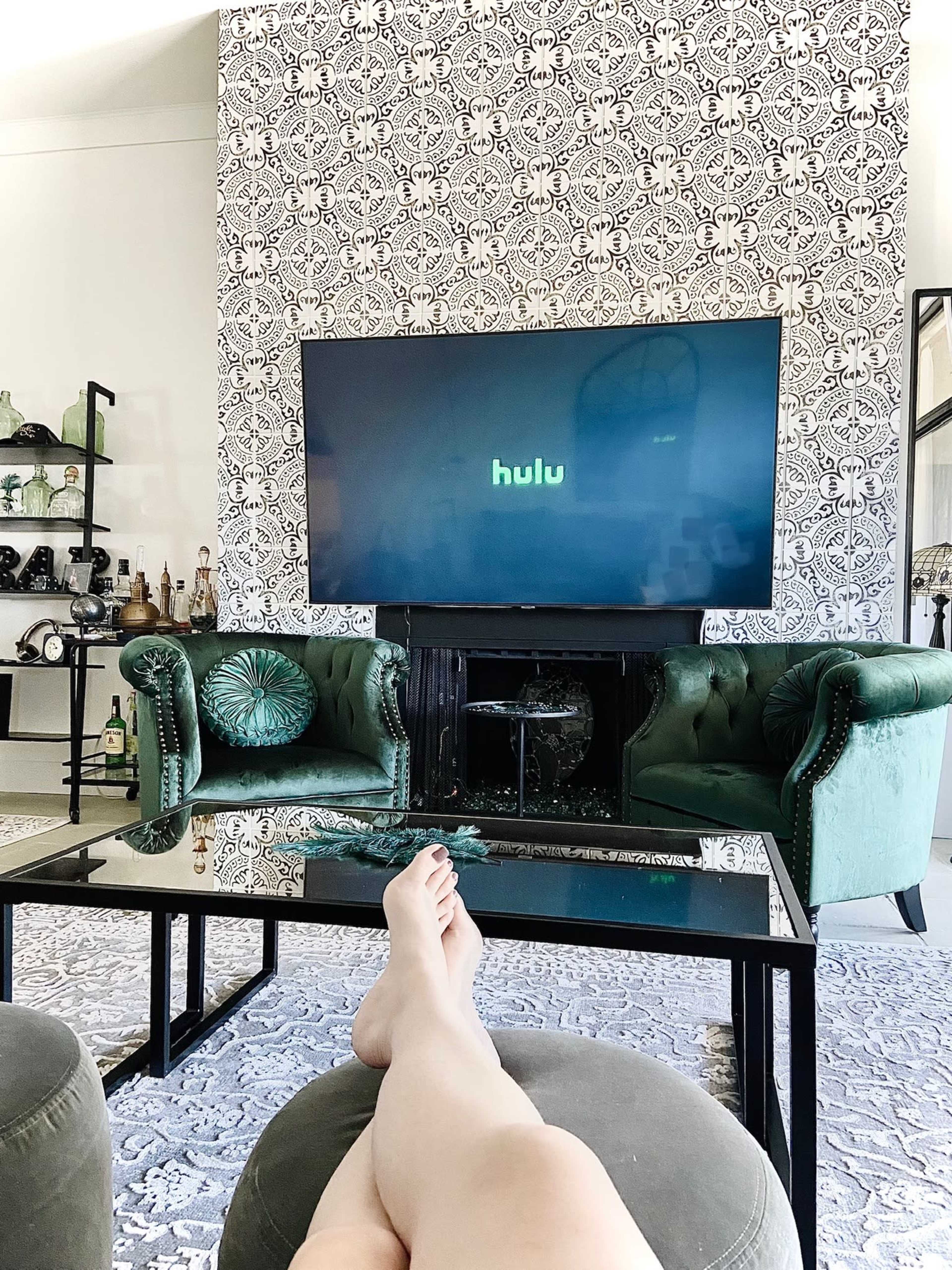 A person is relaxing on a green ottoman in a stylish living room, with a large TV displaying the Hulu logo against a patterned wall.