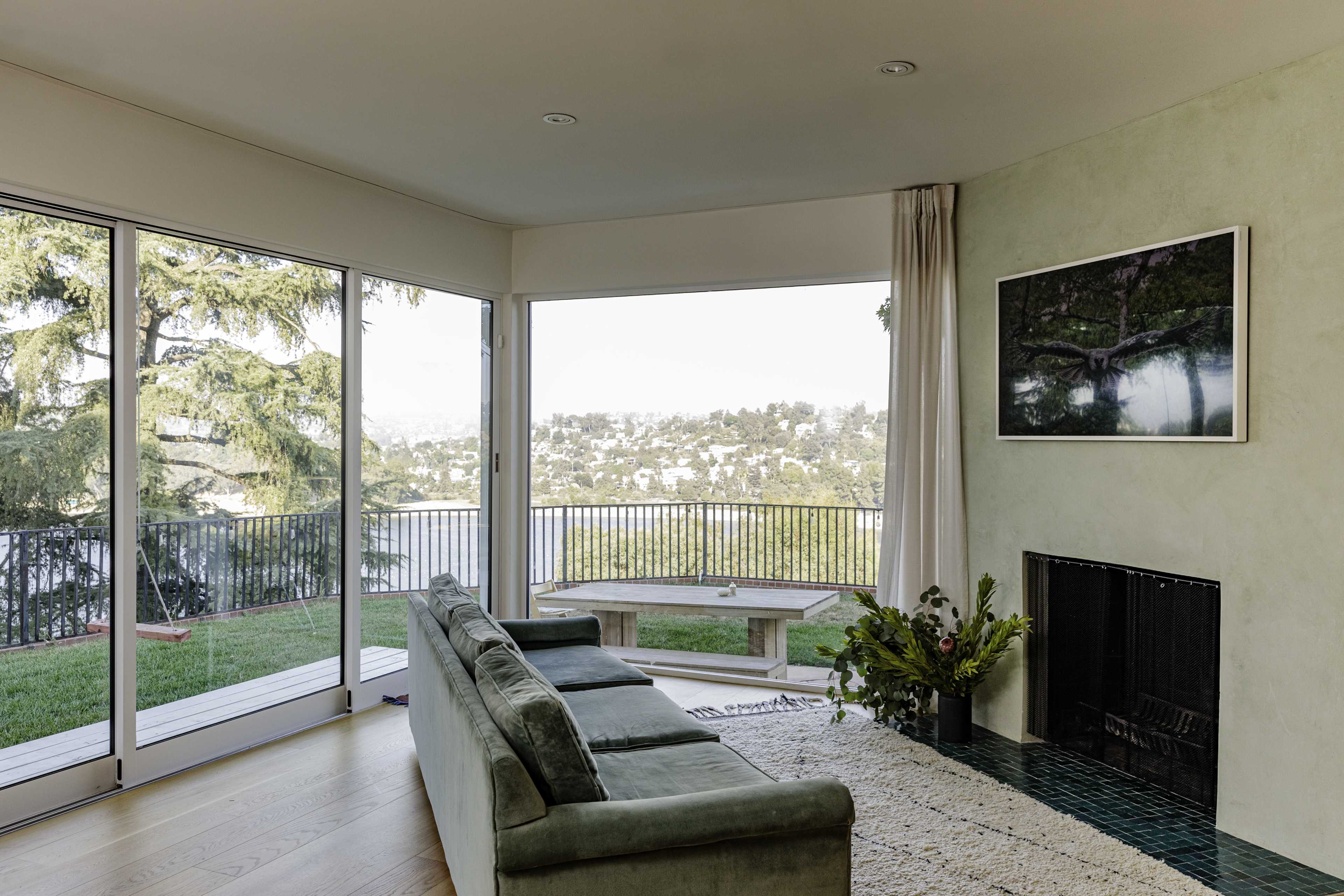 A modern living room features a green sofa, large windows with views of a hillside, and a framed photograph on the wall.