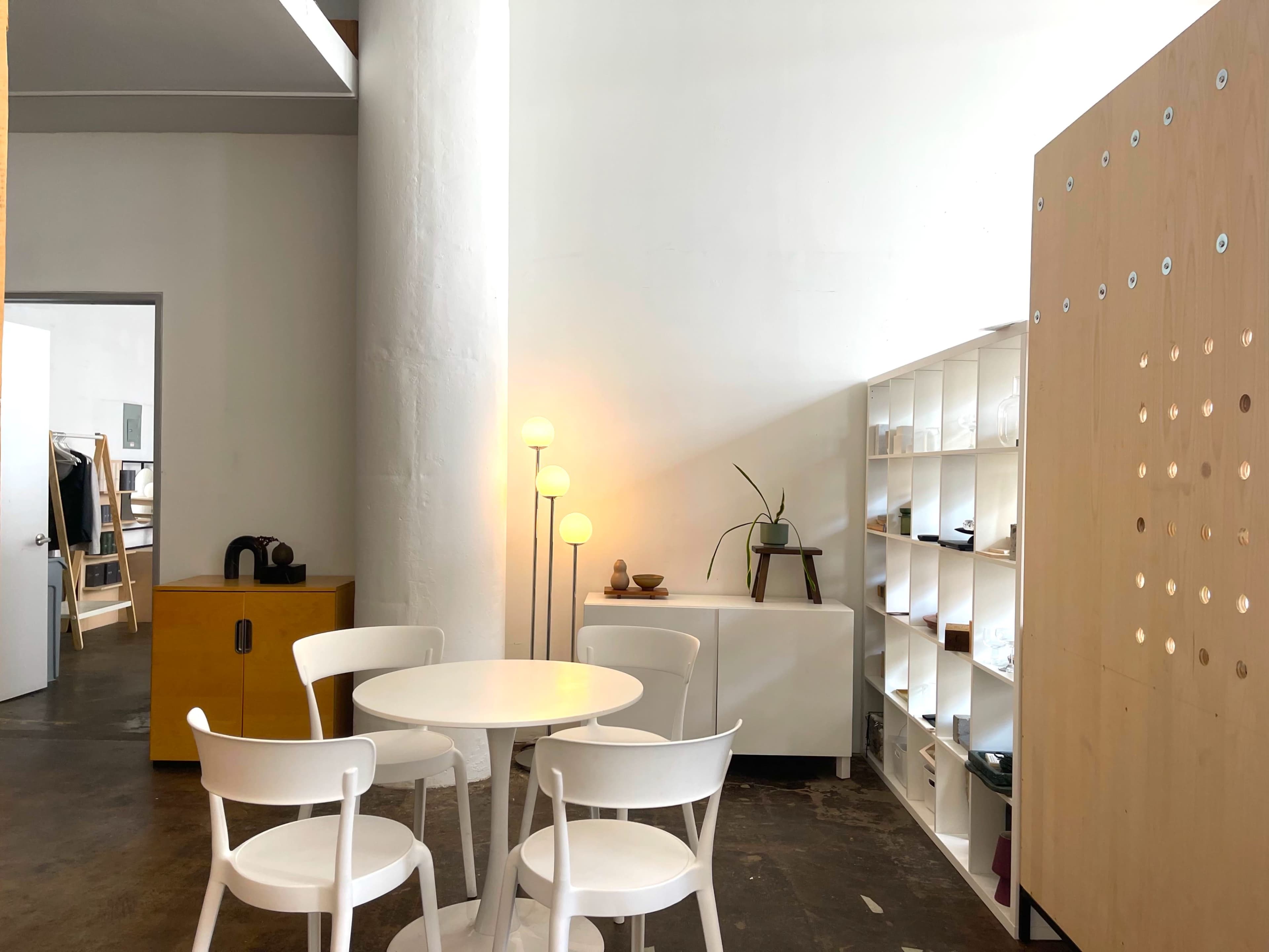 The scene features a small dining area with a round white table and four chairs, alongside a wooden shelving unit and a floor lamp, all within a minimalistic interior.