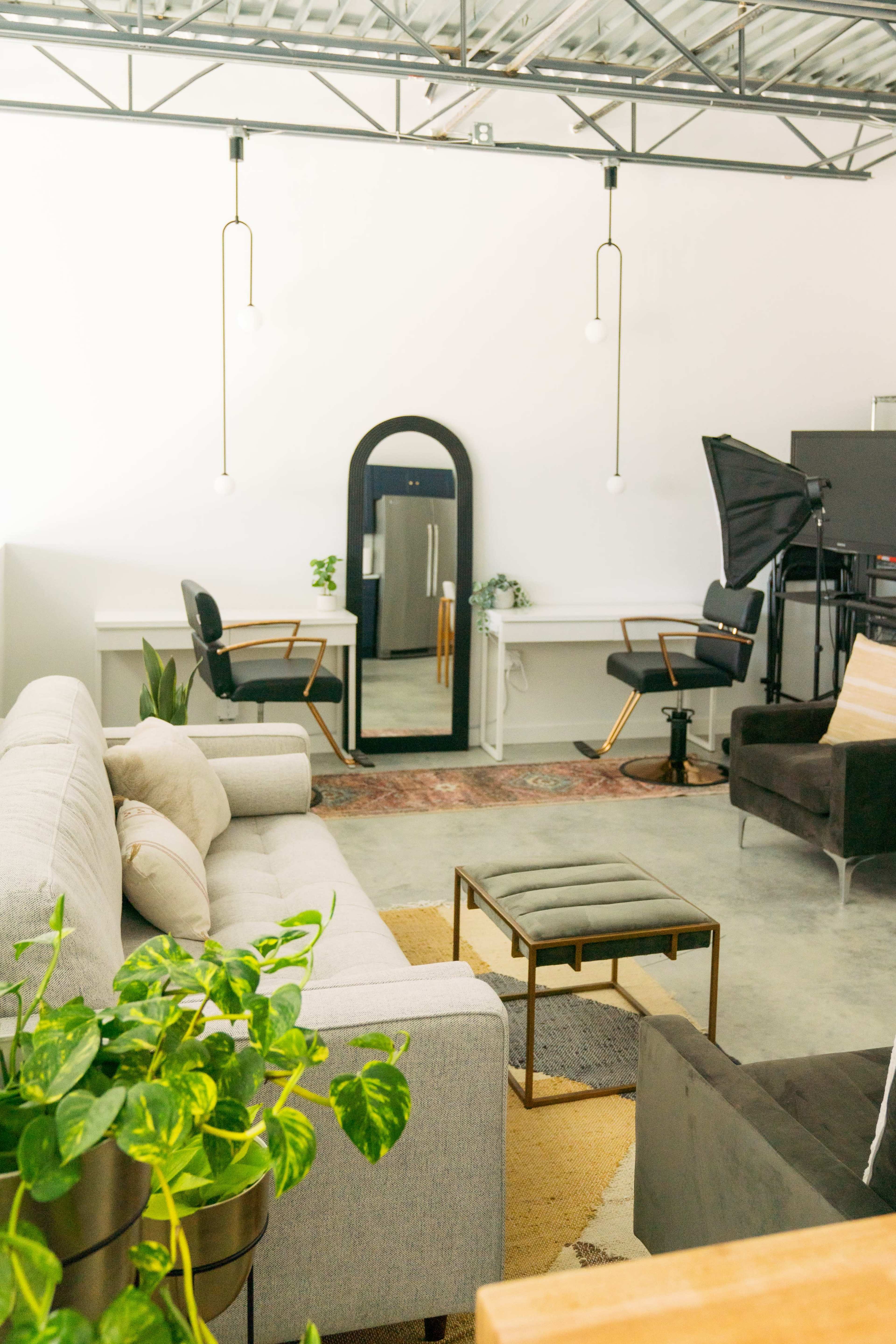 A modern interior space featuring a gray sofa, a coffee table, a large mirror, and workspace chairs, with greenery and soft lighting.
