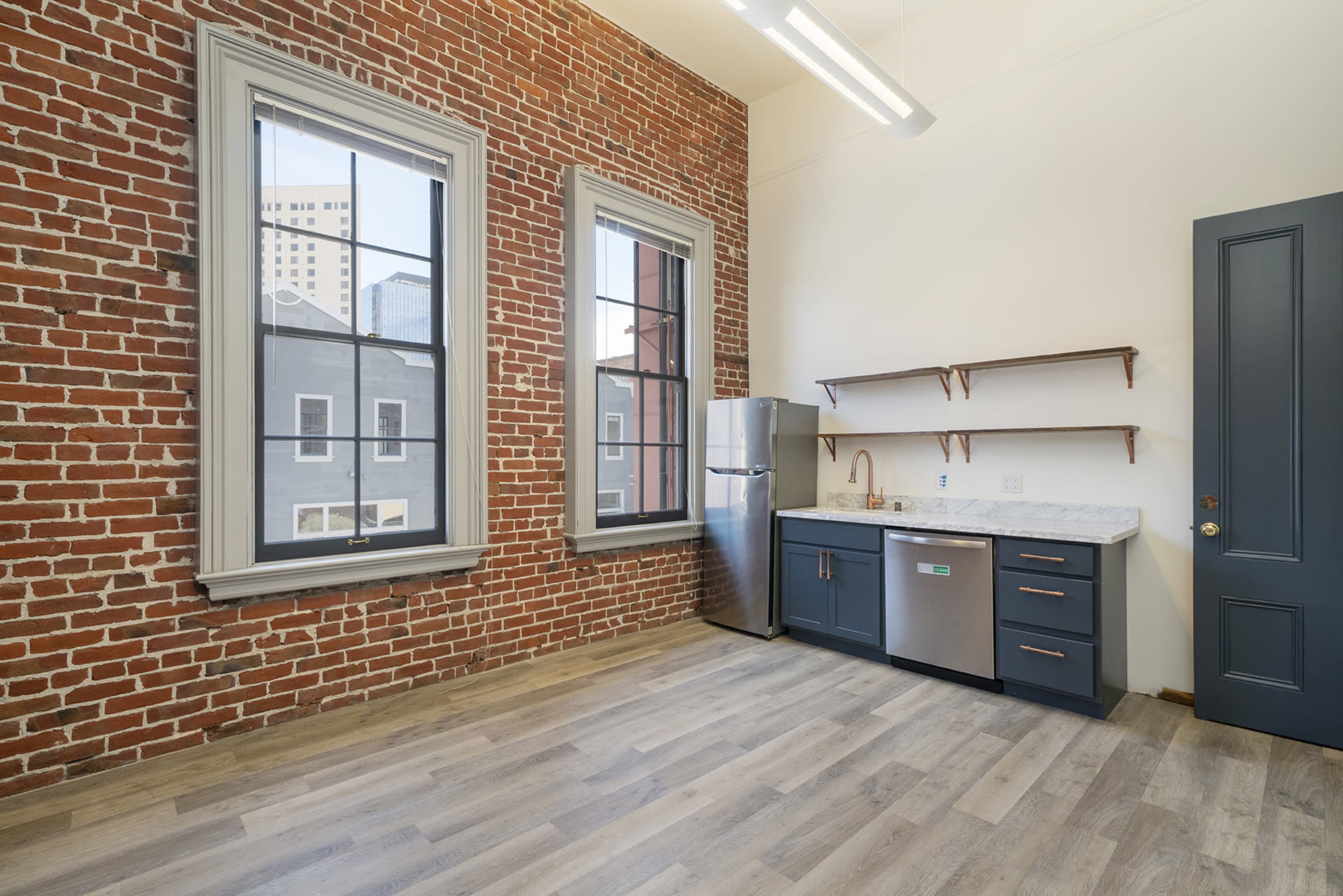 A modern kitchen area in a room with exposed brick walls and large windows, featuring a refrigerator, countertop, and open shelving.