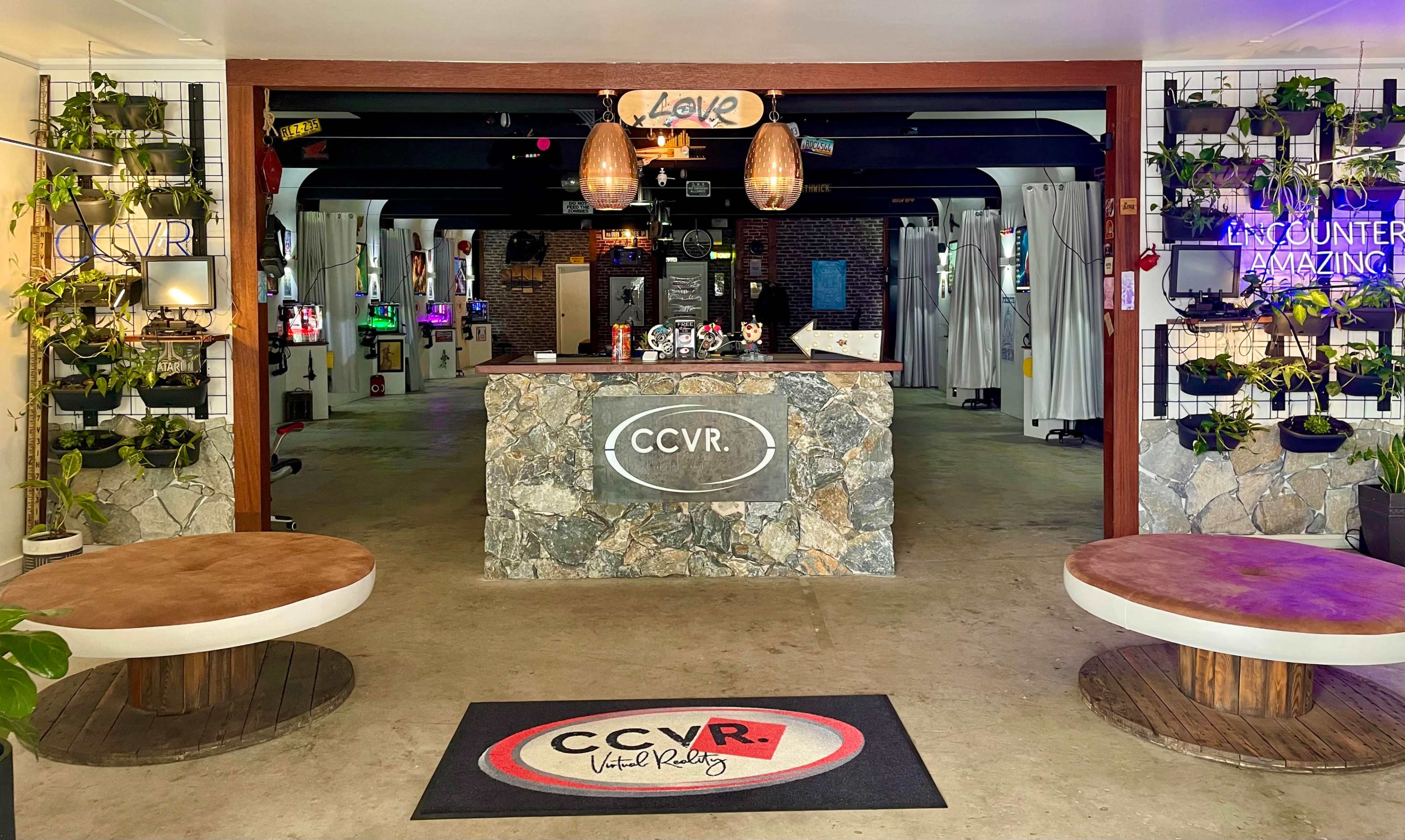 A modern reception area for a virtual reality venue, featuring a stone counter, potted plants, and gaming stations in the background.