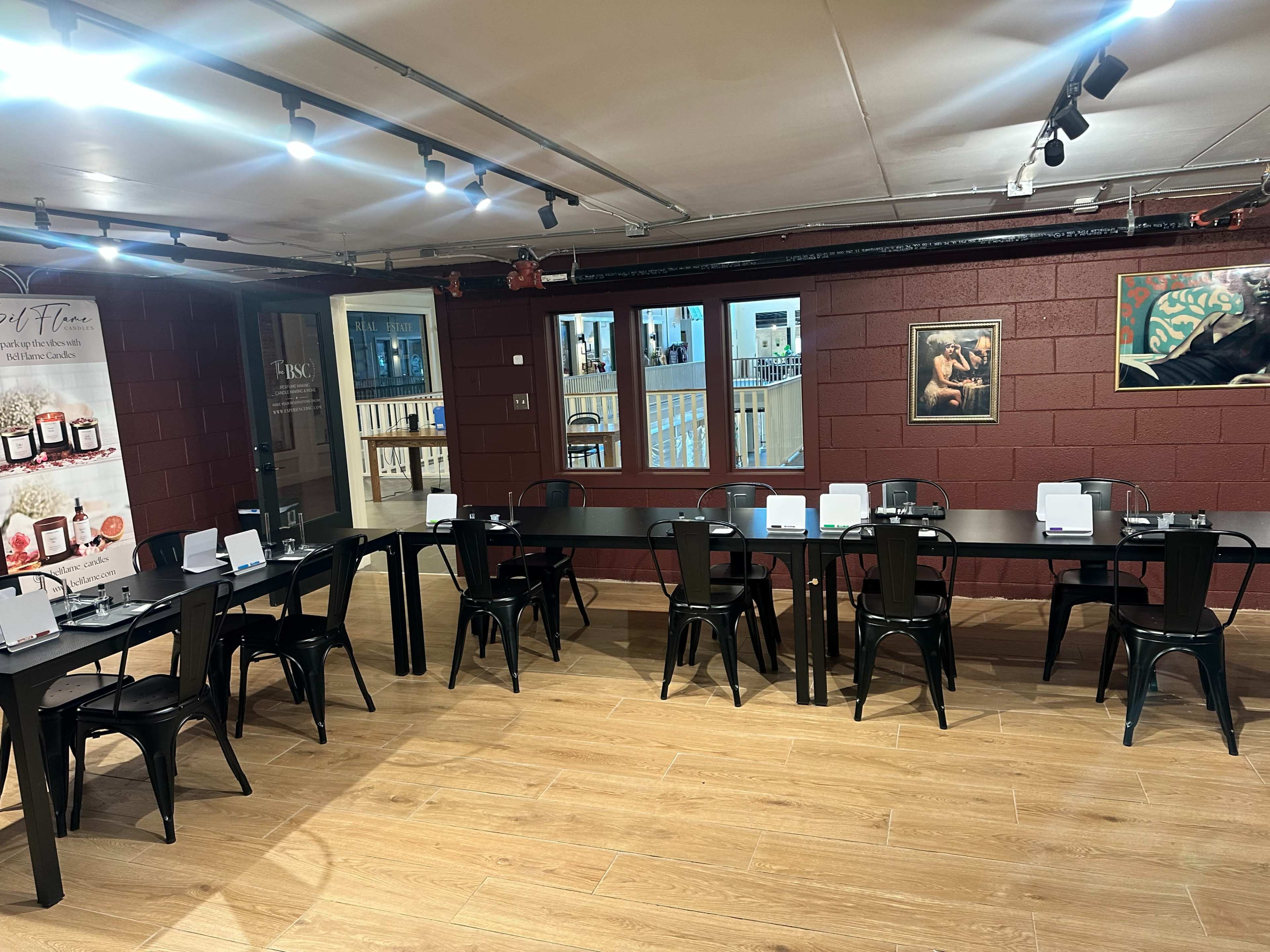 The image shows a dimly lit dining area with black tables and chairs arranged in two rows, and a wall featuring artwork and promotional displays.