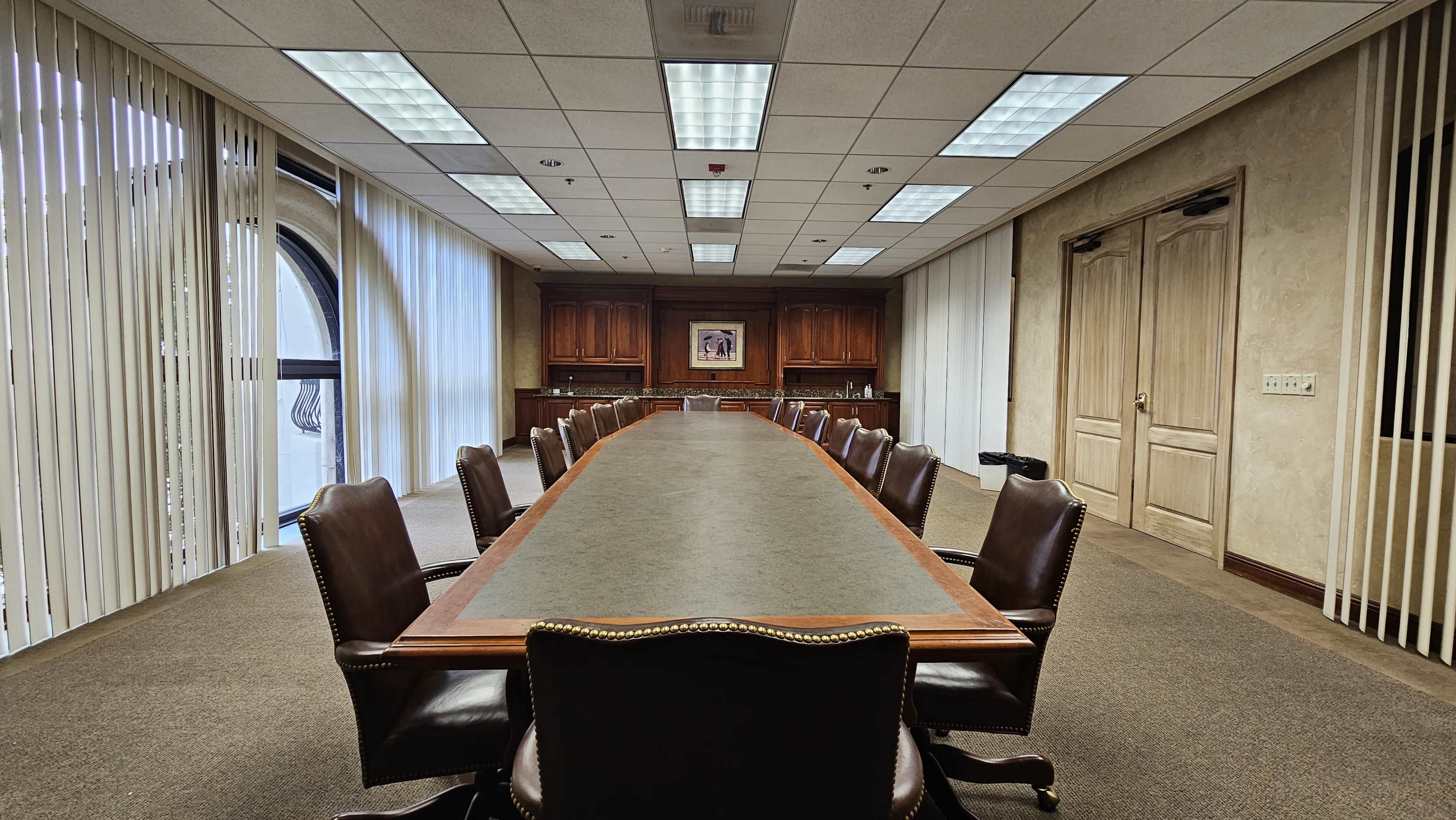 A long, rectangular conference table is set in a well-lit meeting room with wooden cabinetry and large windows covered by vertical blinds.