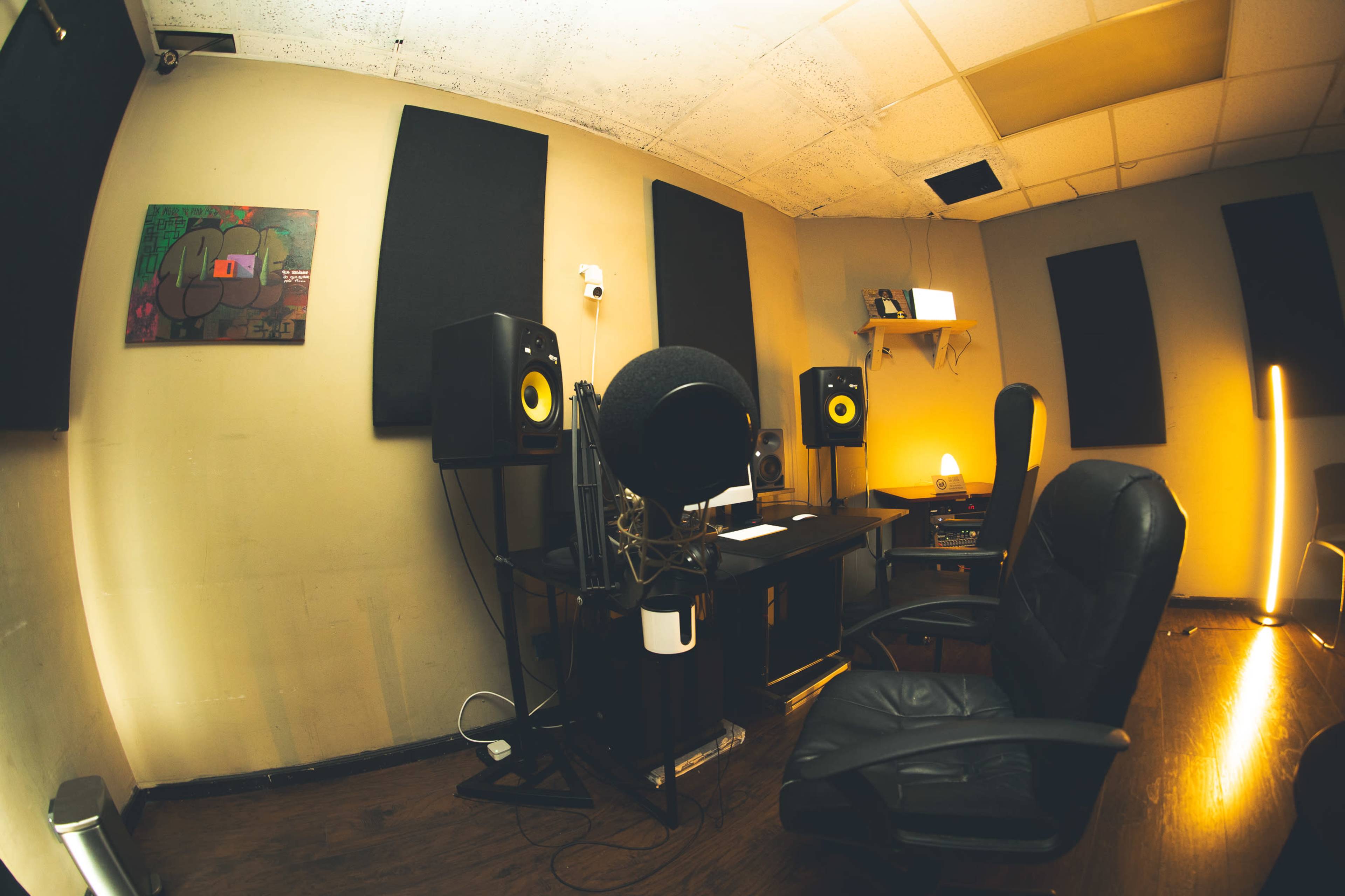 The image shows a small recording studio with a microphone setup, audio monitors, and a desk in a room with soundproofing panels on the walls.