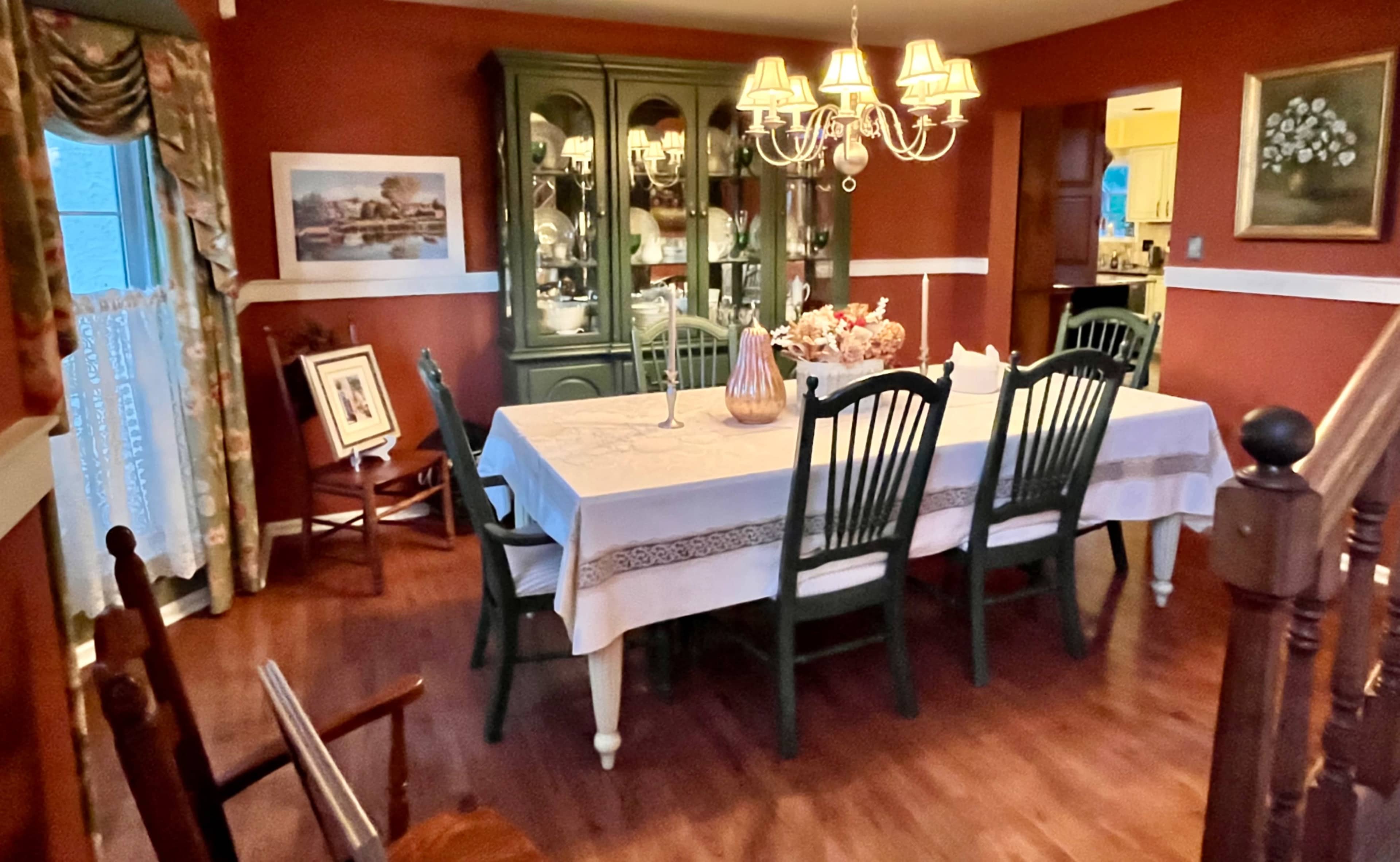 A large dining table with green chairs and a chandelier is set in a warmly decorated dining room featuring wooden flooring and walls painted in a deep color.