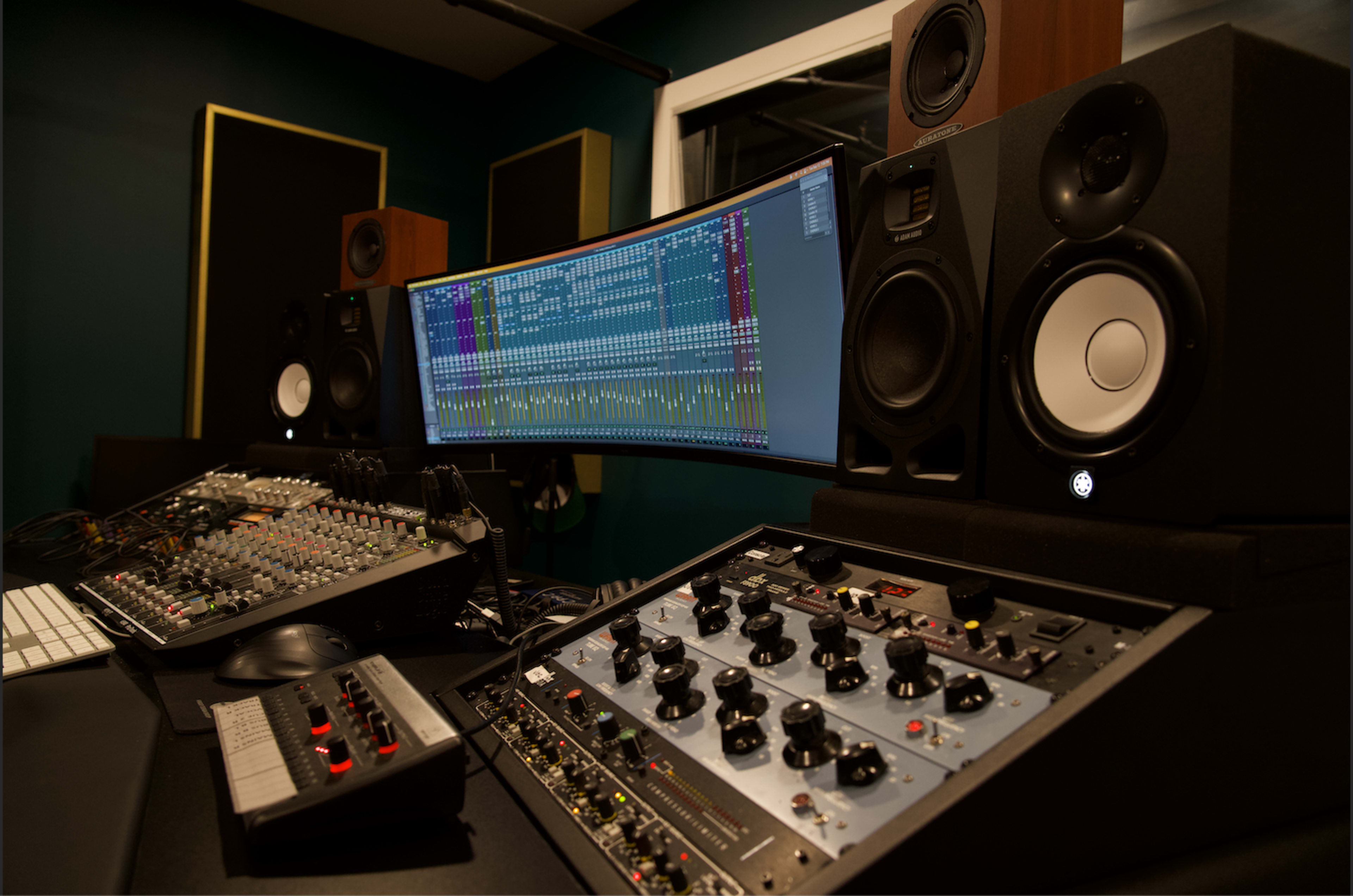 A music production studio with a curved monitor displaying audio tracks, a mixing console, and studio speakers.