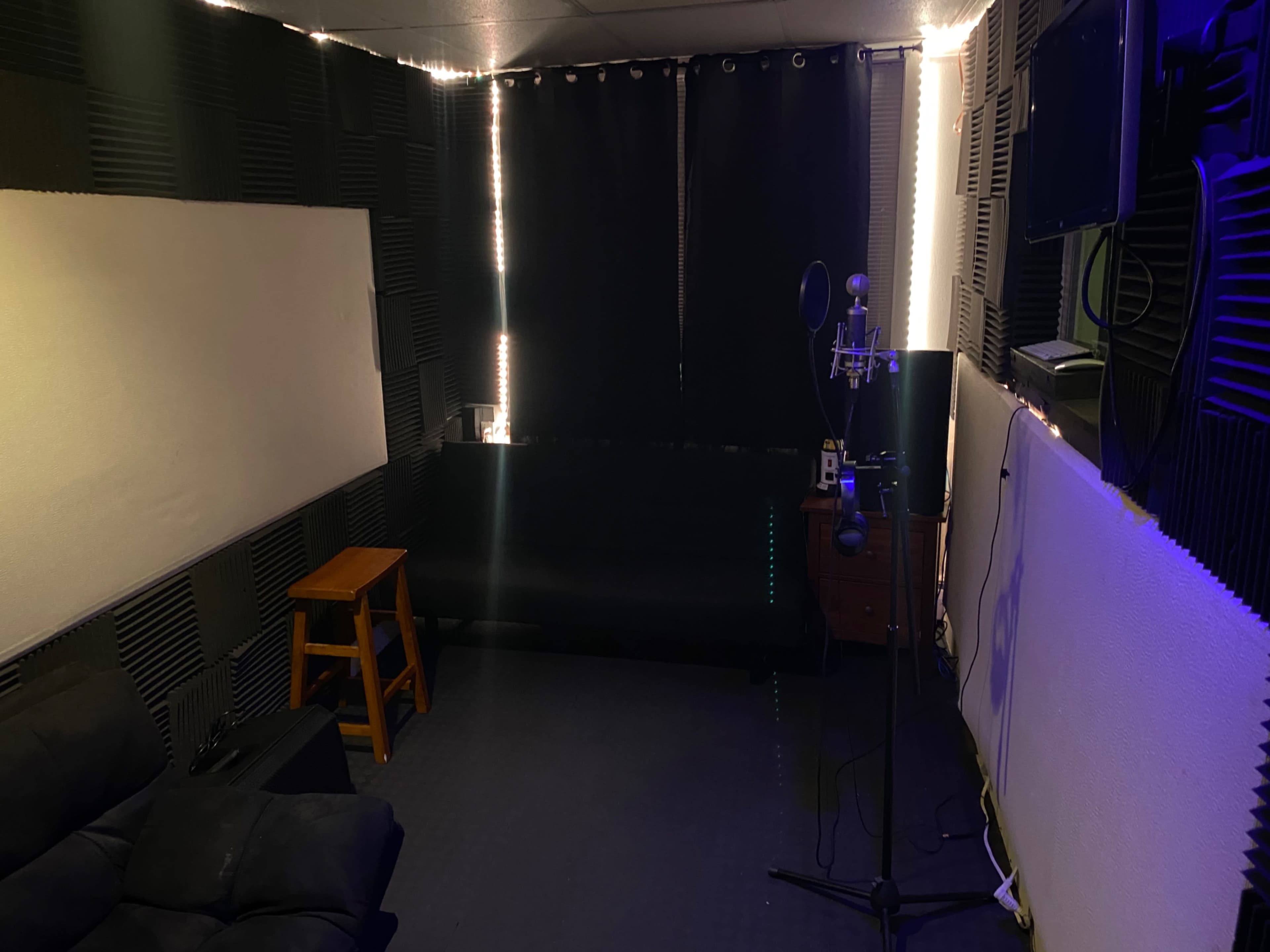 A small recording studio with soundproofing panels, a microphone on a stand, a couch, and a stool against dark curtains.