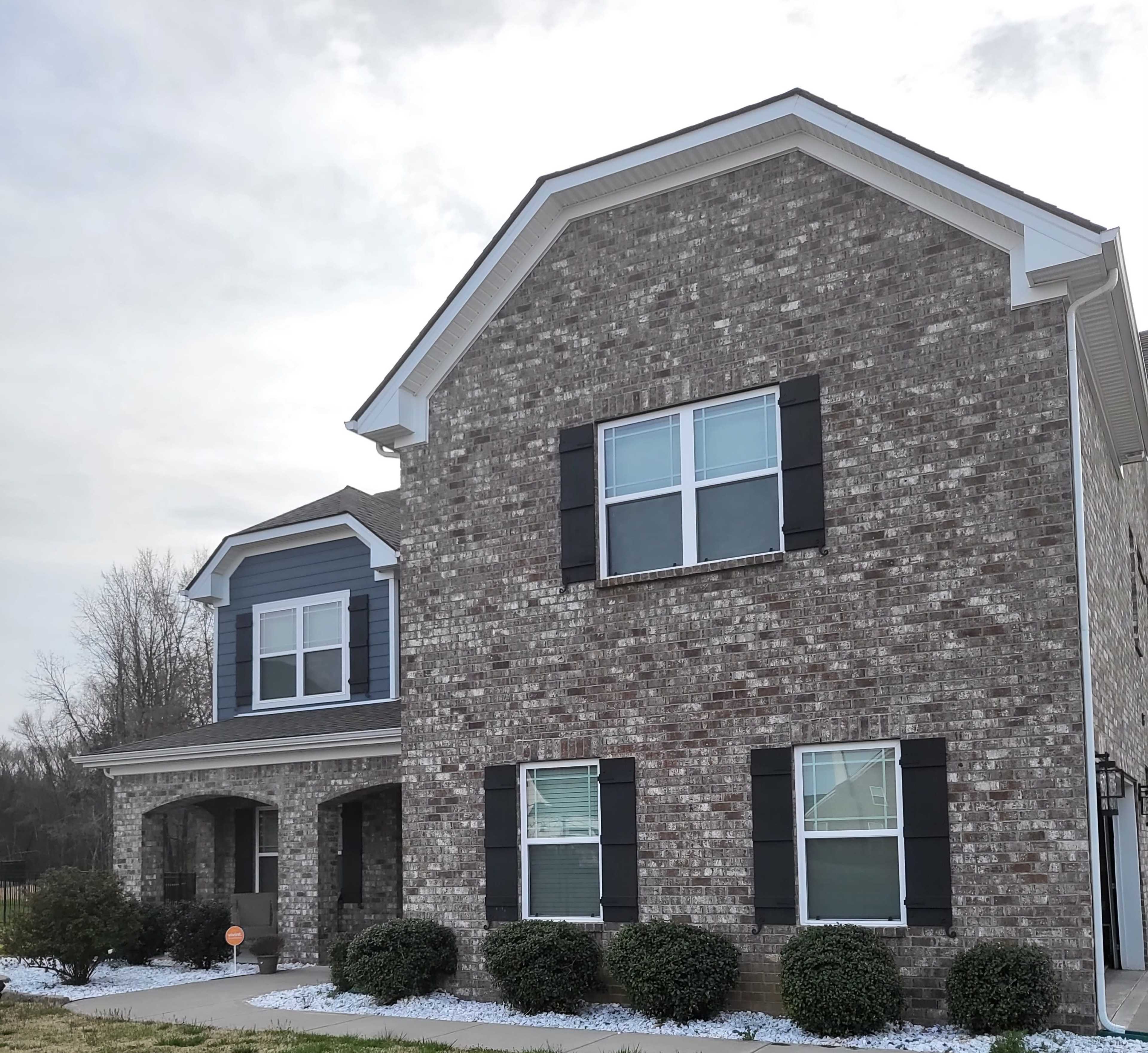 A two-story brick house with a combination of brown and blue siding, featuring black shutters and landscaped shrubbery in front.