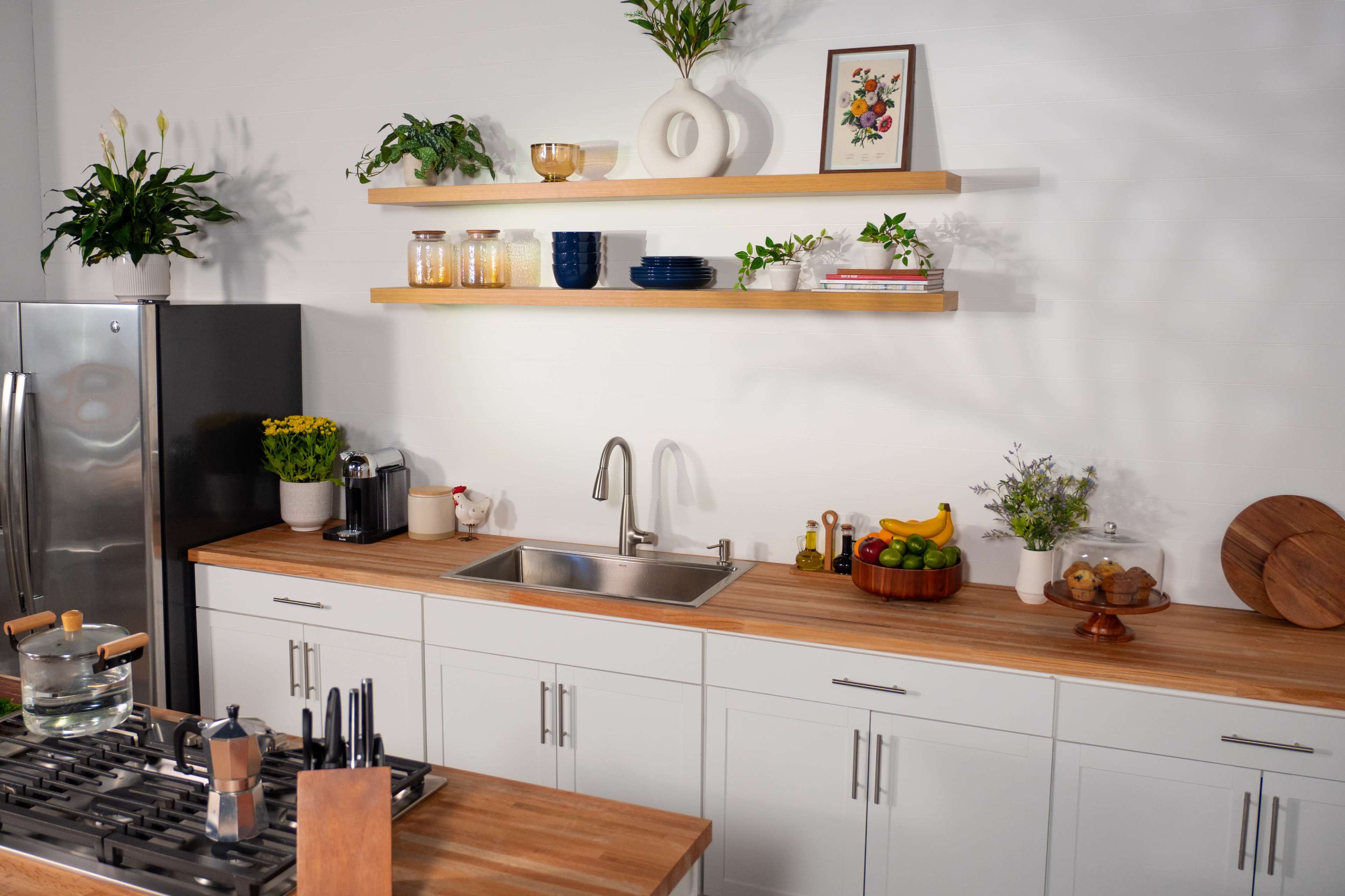 A modern kitchen features a stainless steel refrigerator, a double sink, wooden shelves with decorative items, and a countertop with various kitchen accessories and fruits.