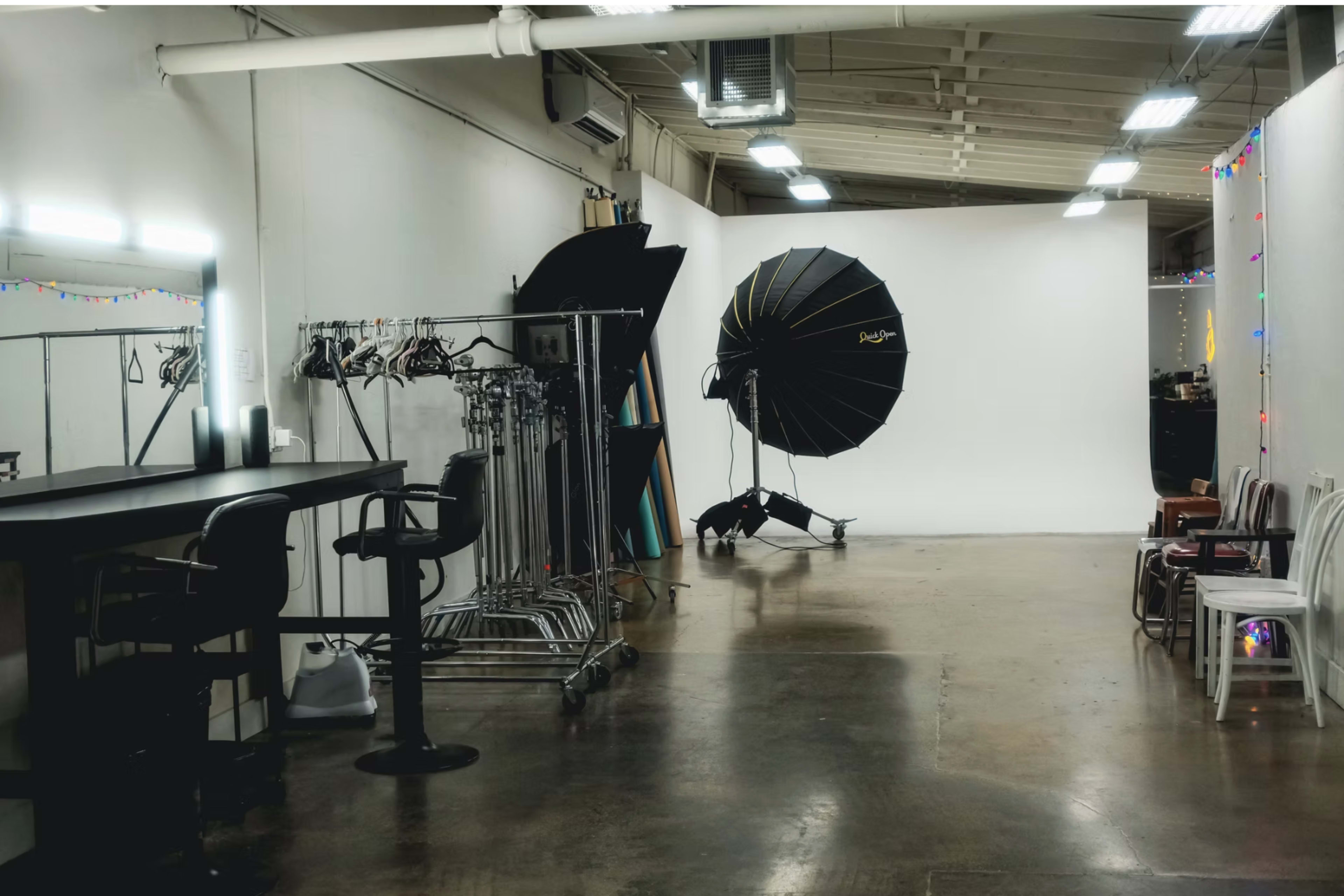 The image shows a photography studio with a large backdrop, lighting equipment, and clothing racks along the wall.