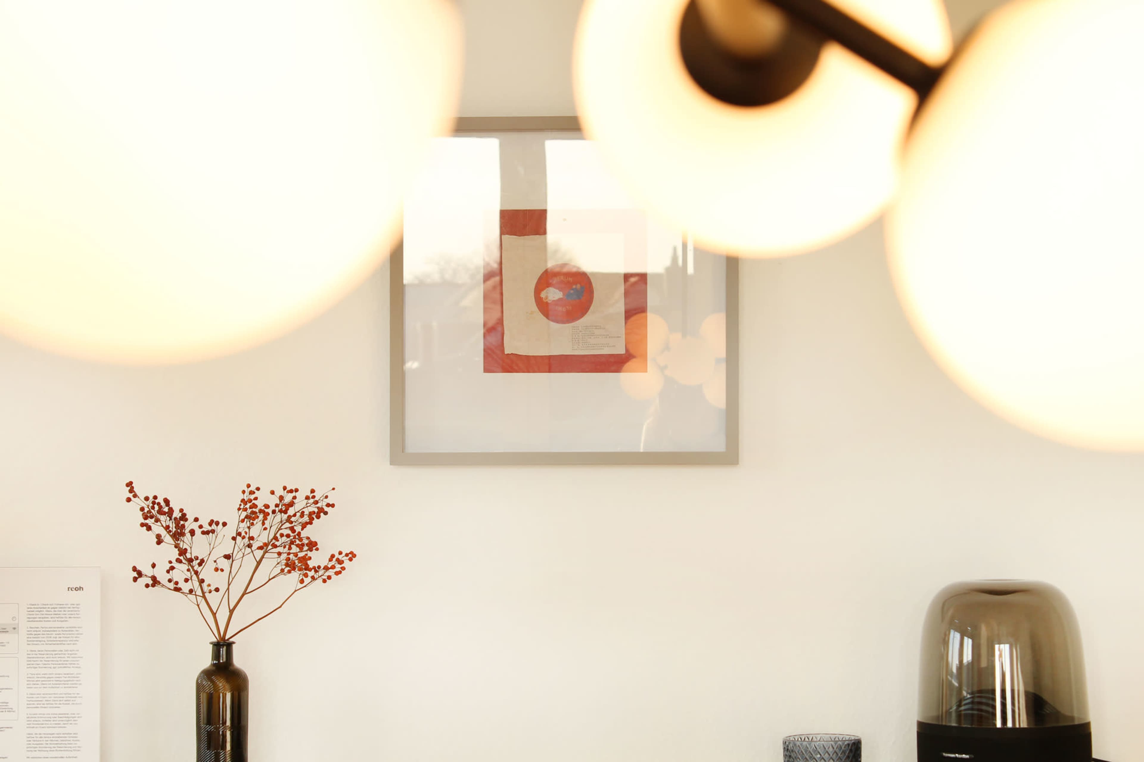 A modern interior featuring a framed artwork on a wall, with a decorative vase and a kitchen appliance visible on a countertop, partially obscured by lighting fixtures.