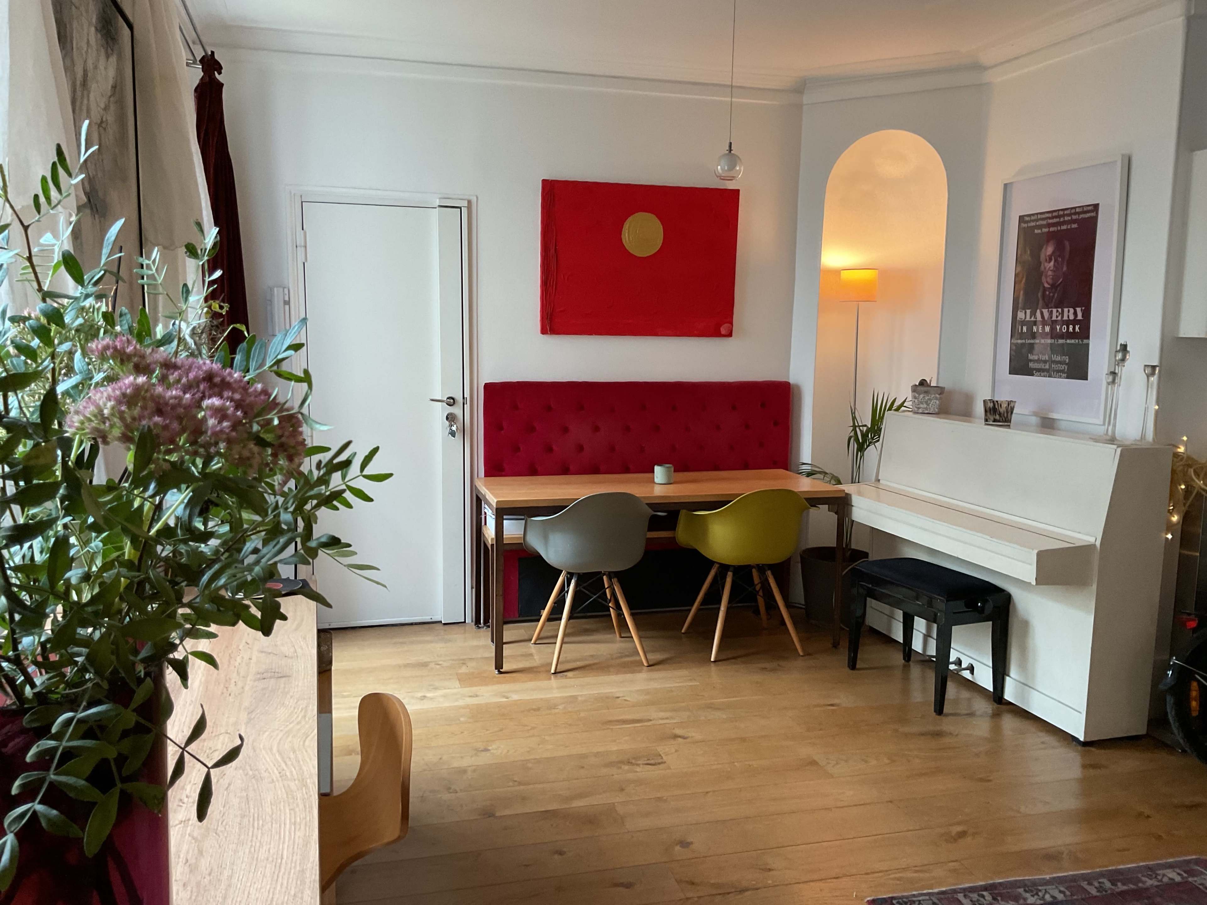 A cozy dining area features a red tufted bench along a white wall, a wooden table with colorful chairs, and a piano in a well-lit space.