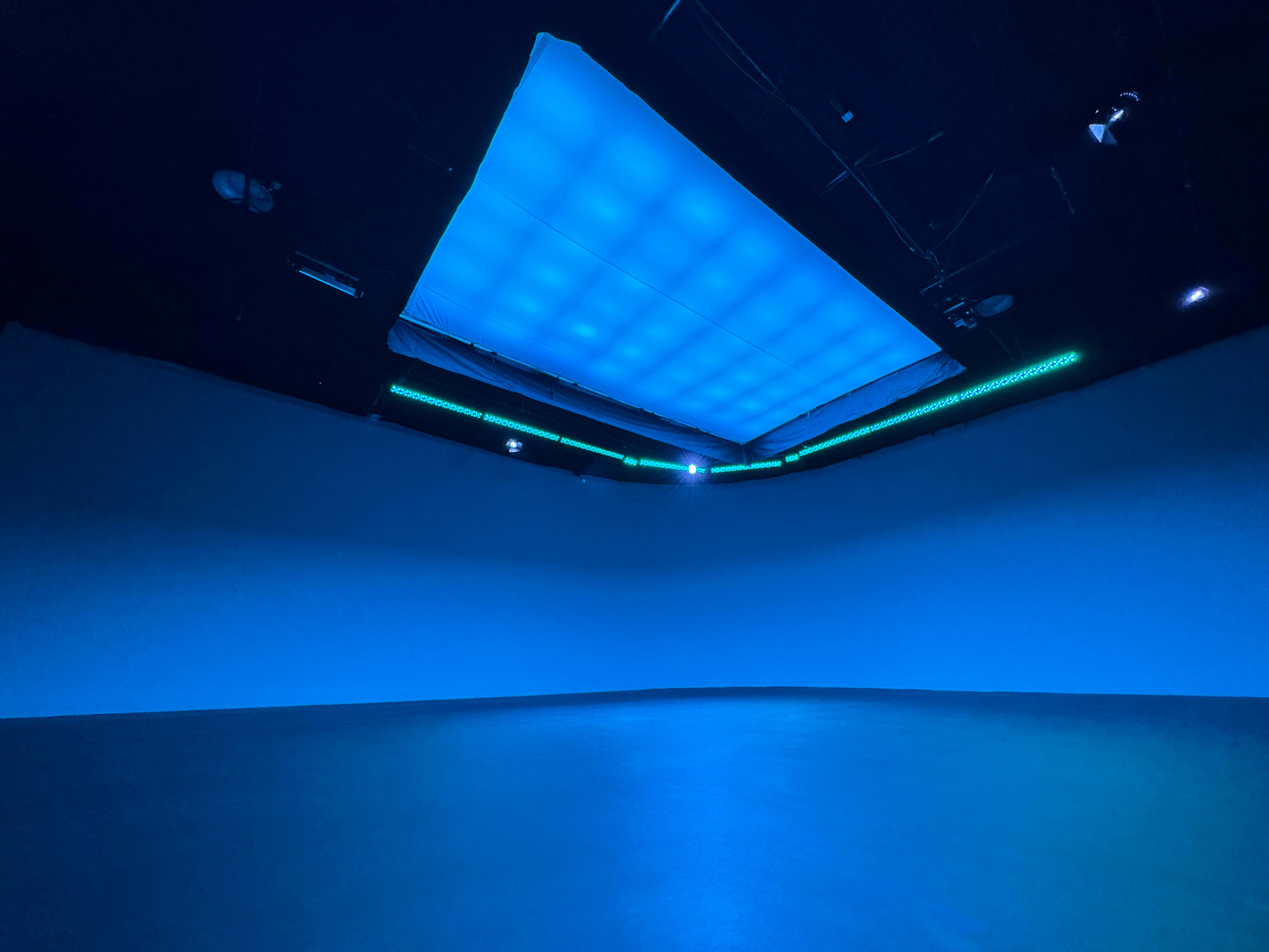A dark exhibition space with a large, illuminated blue panel on the ceiling and green lights along the upper walls.