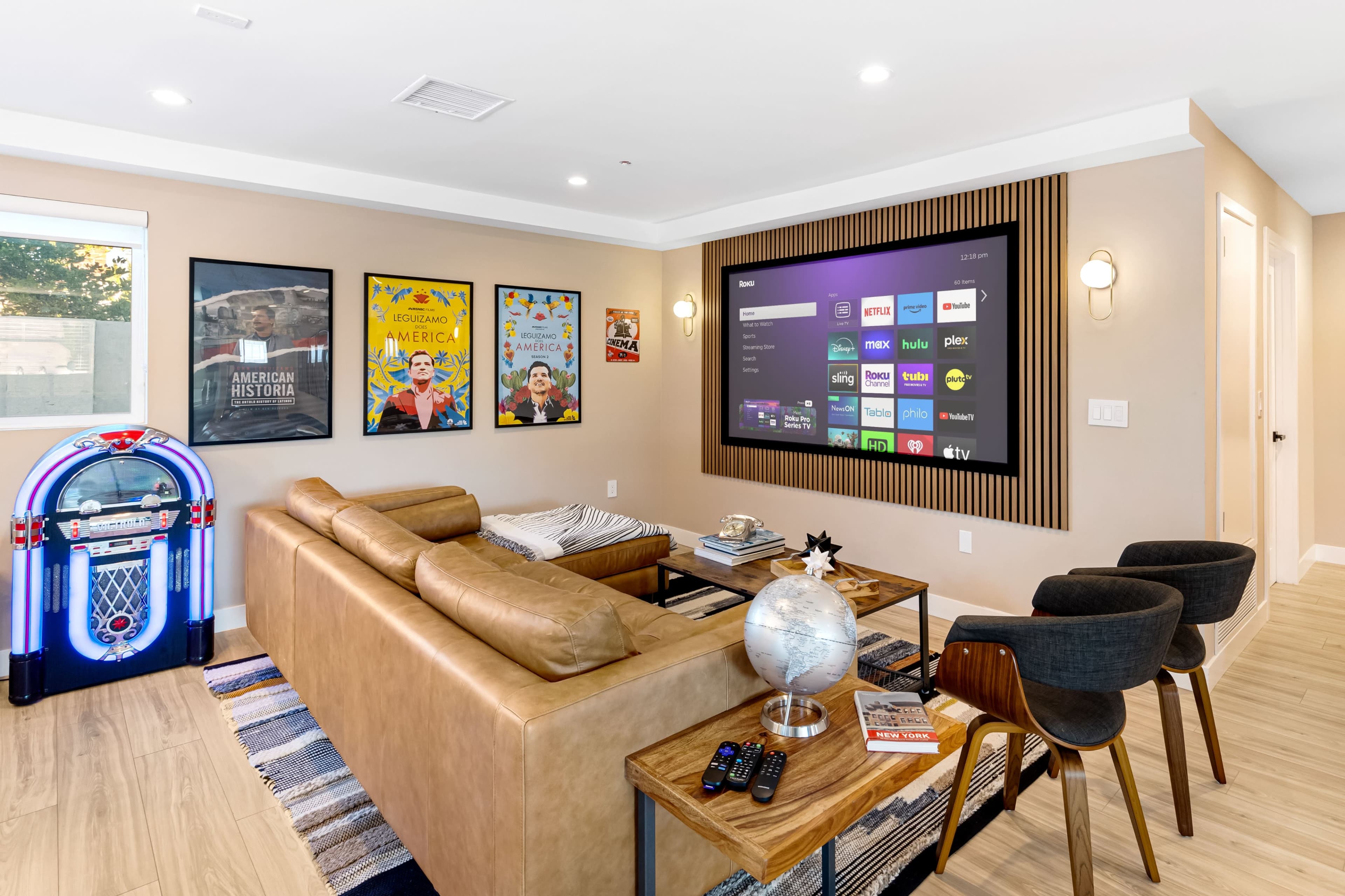 The image shows a modern living room with a large wall-mounted television, a leather sofa, an arcade-style jukebox, and various framed posters on the walls.