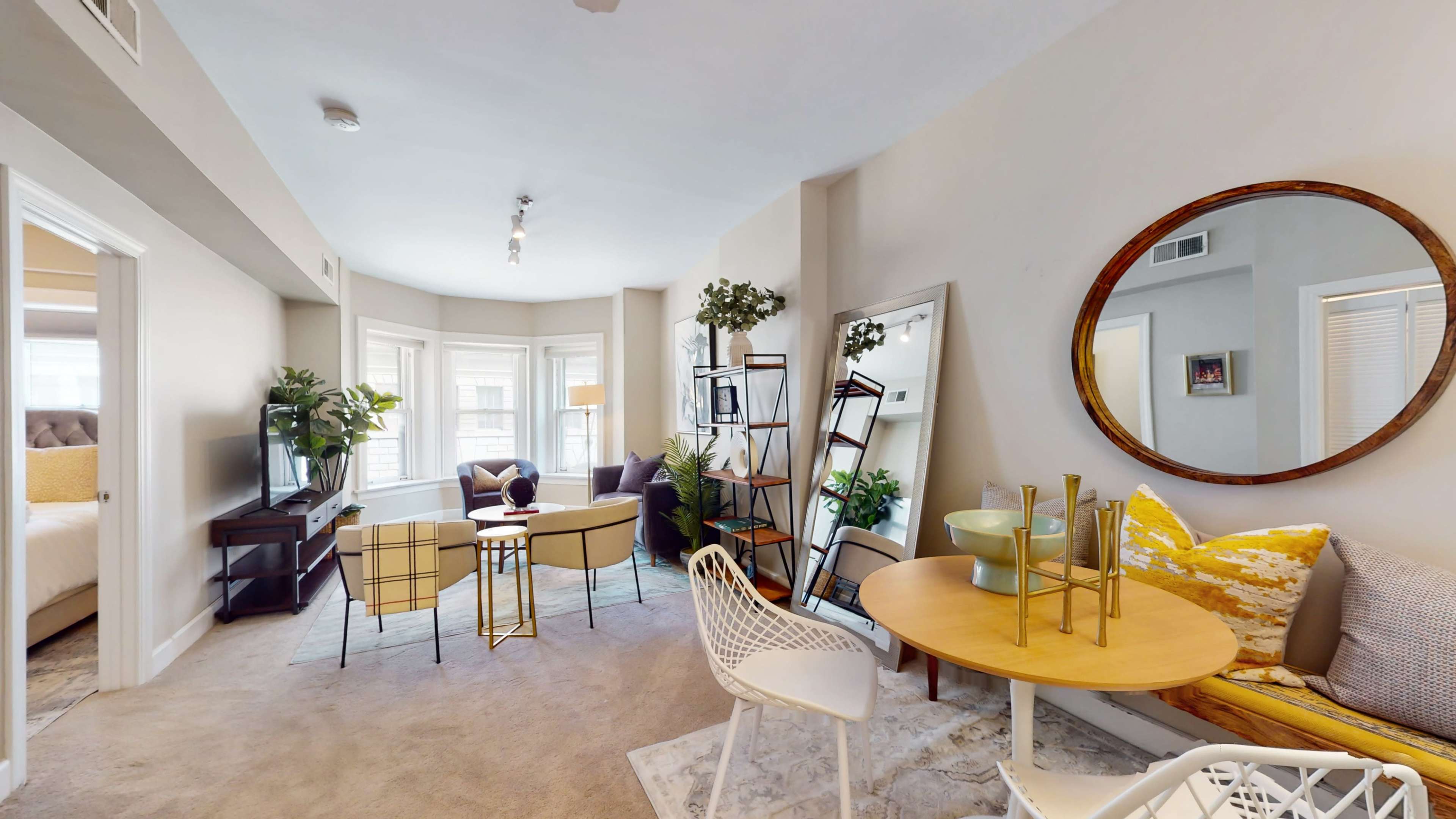 The image depicts a well-furnished living space featuring a seating area with a round table, a television stand, and large windows with natural light, surrounded by decorative plants and a mirror.