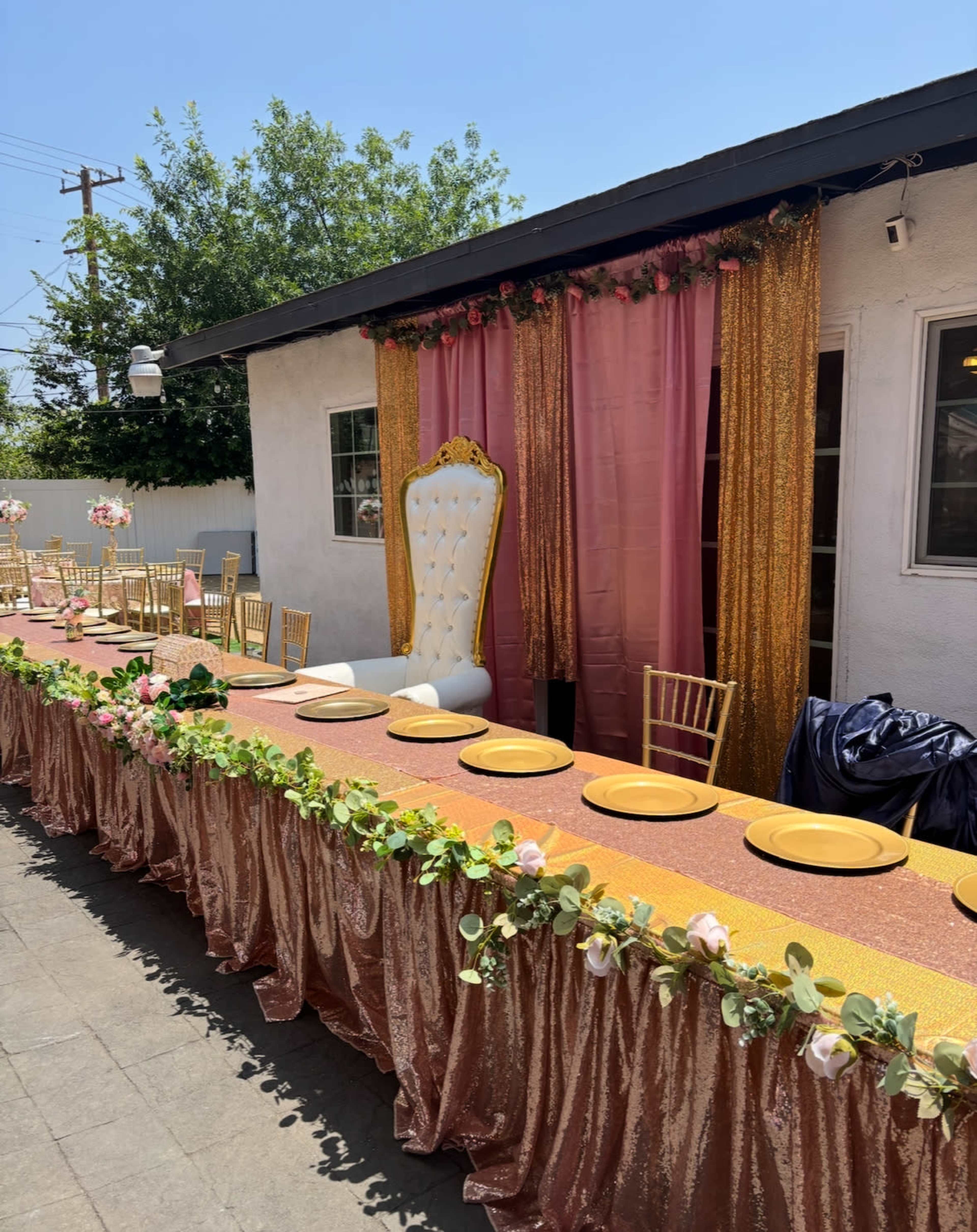 The outdoor event setting features a long banquet table adorned with gold plates and floral decorations, alongside a large white throne chair against a backdrop of decorative curtains.