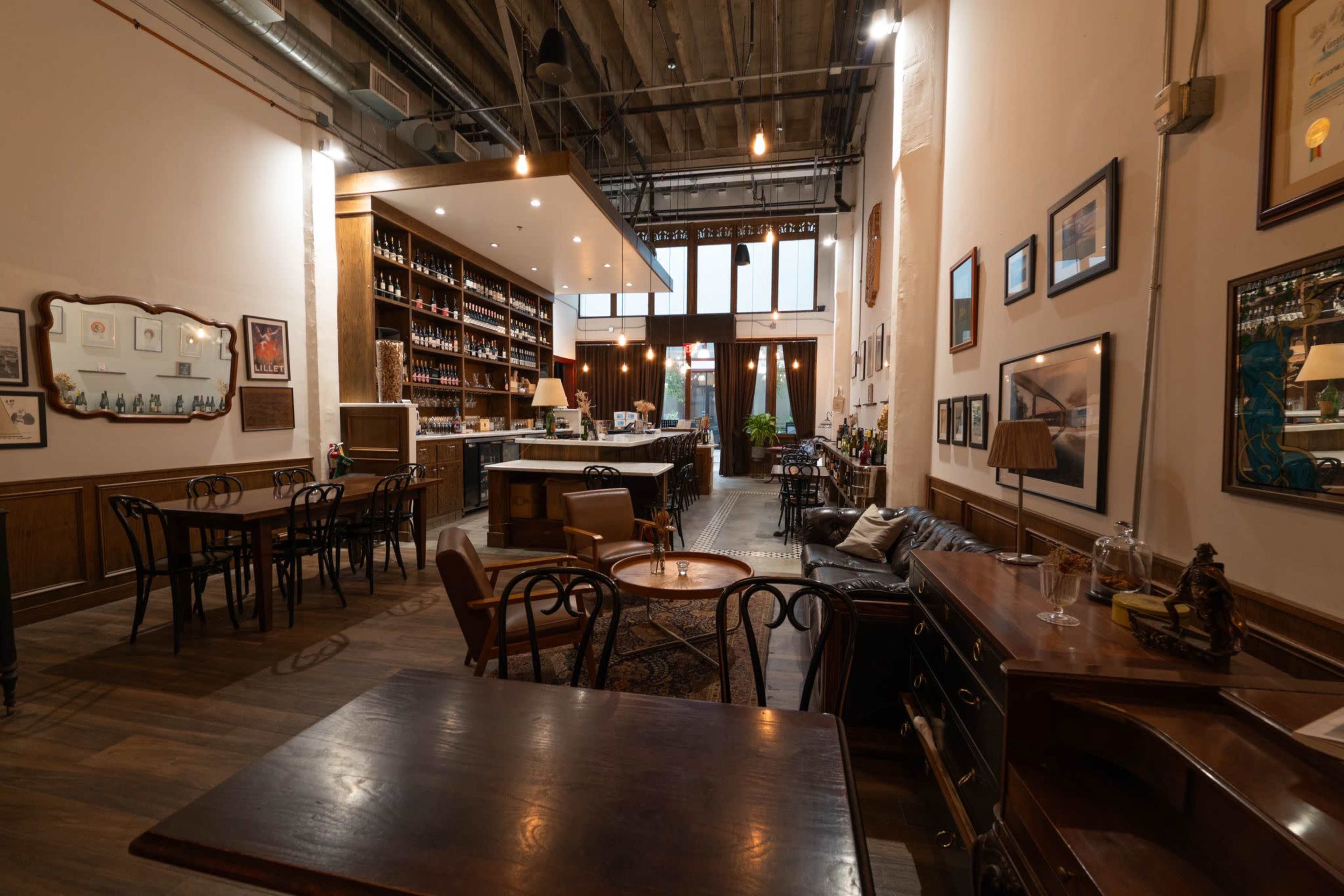 The image shows a cozy cafe interior featuring wooden furniture, shelves filled with bottles, and framed artwork on the walls.