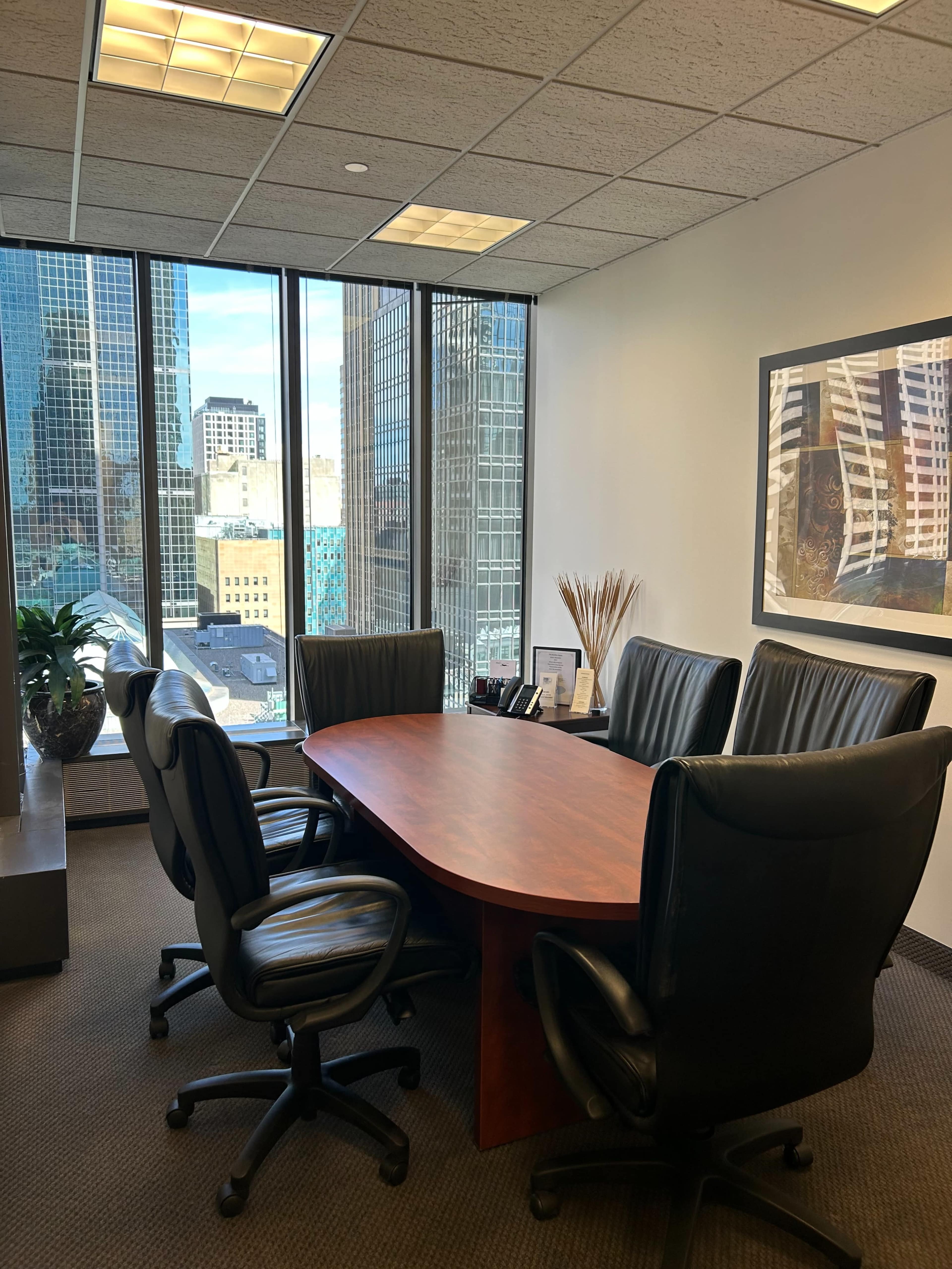 A conference room features a large oval table surrounded by six black leather chairs, with large windows offering a view of skyscrapers outside.