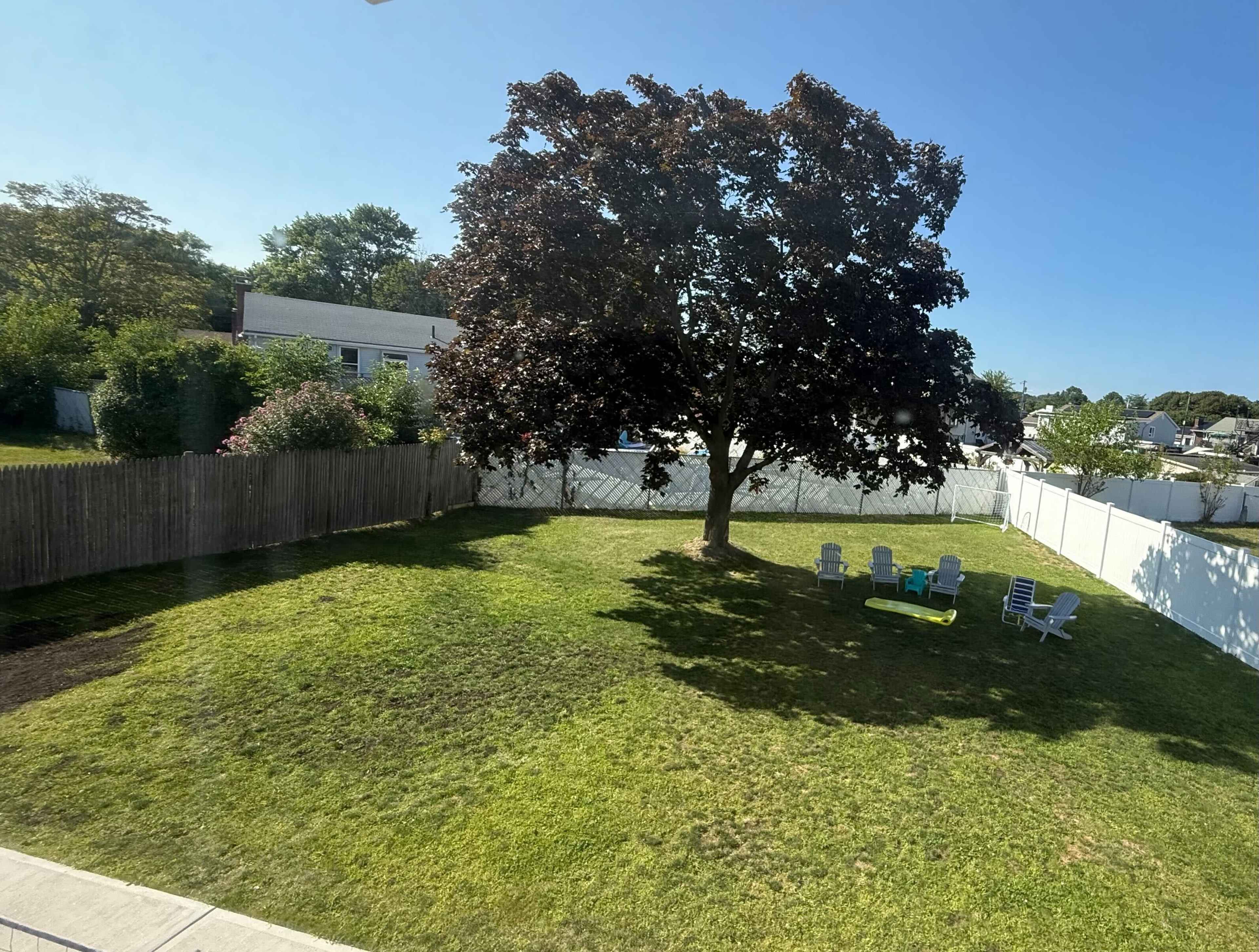 A large tree provides shade over a grassy yard with a few chairs arranged around a small table.