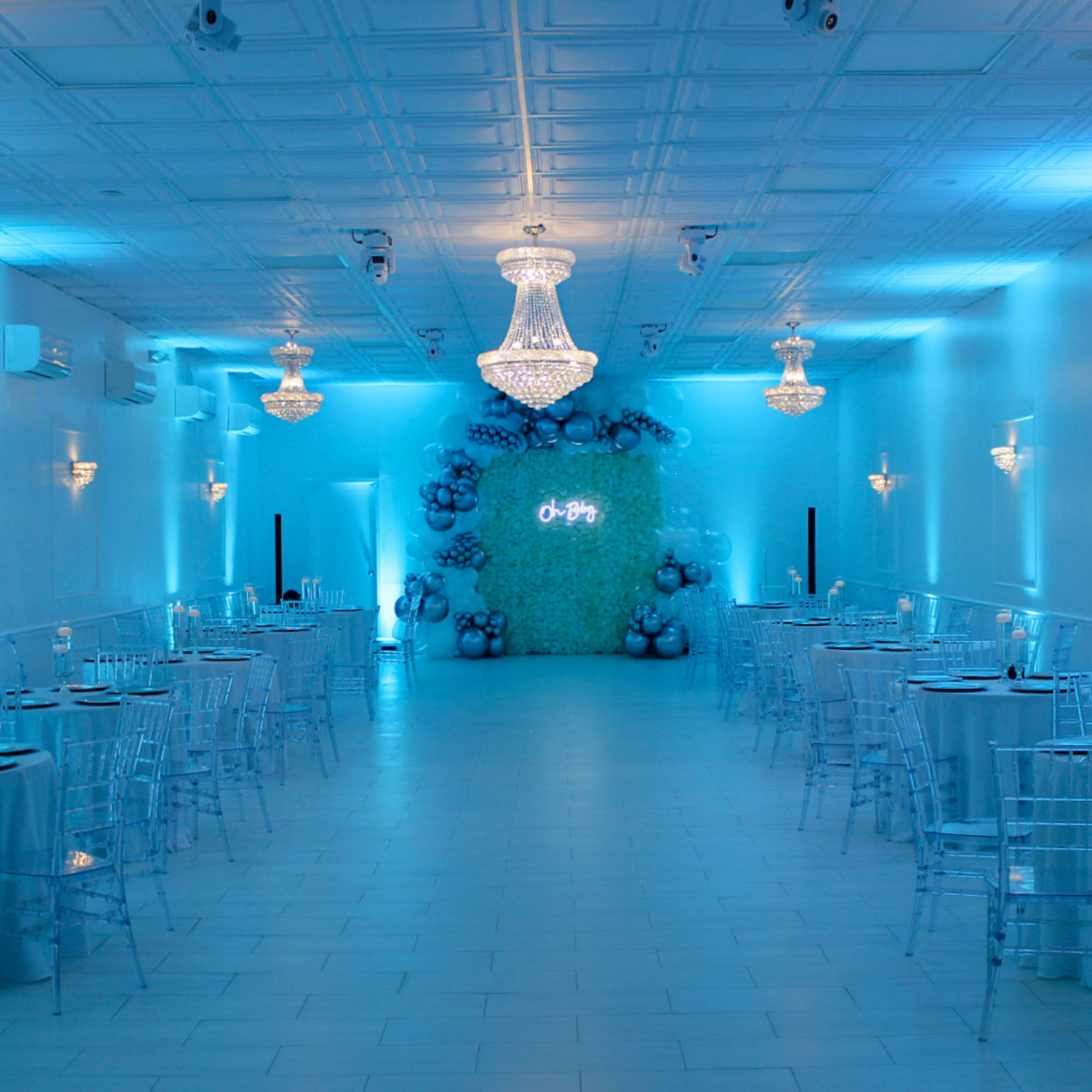 A spacious event venue adorned with blue lighting, elegant chandeliers, and tables set with chairs, featuring a decorative balloon backdrop.