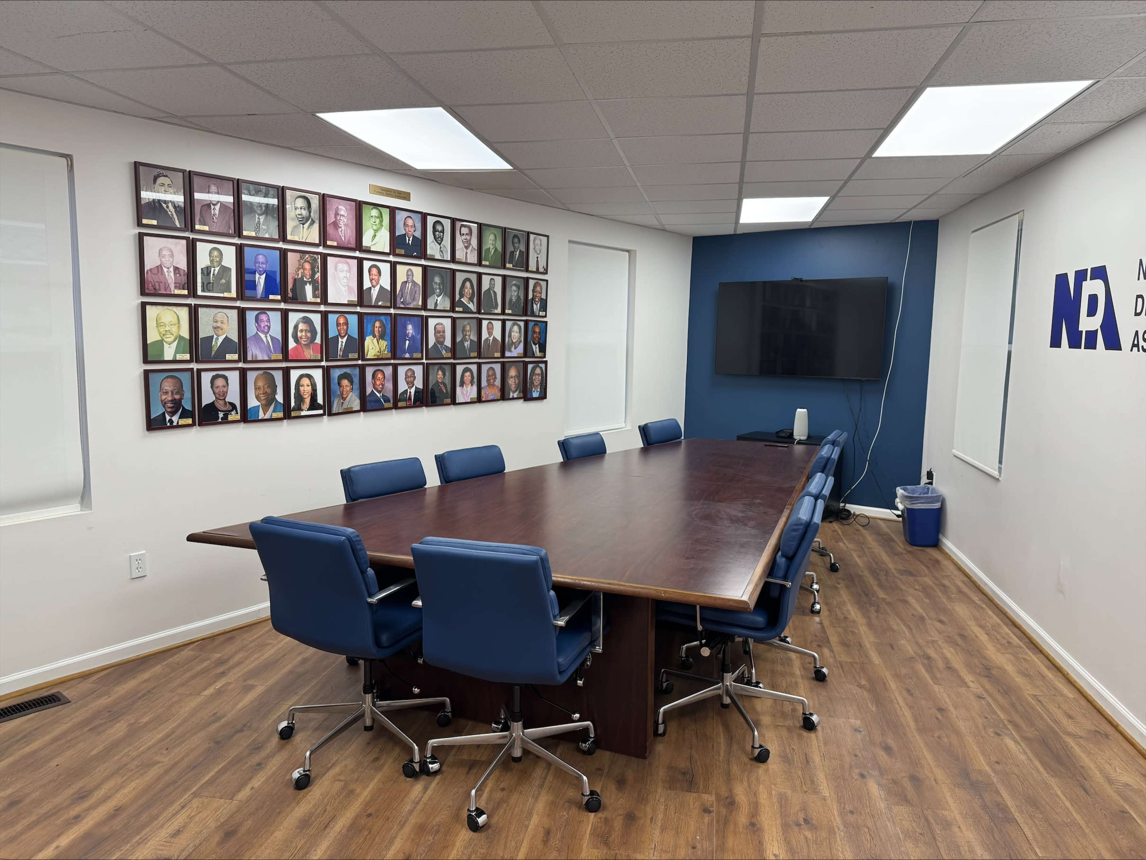 The image shows a conference room with a large wooden table surrounded by blue rolling chairs, and a wall featuring a grid of framed portraits.