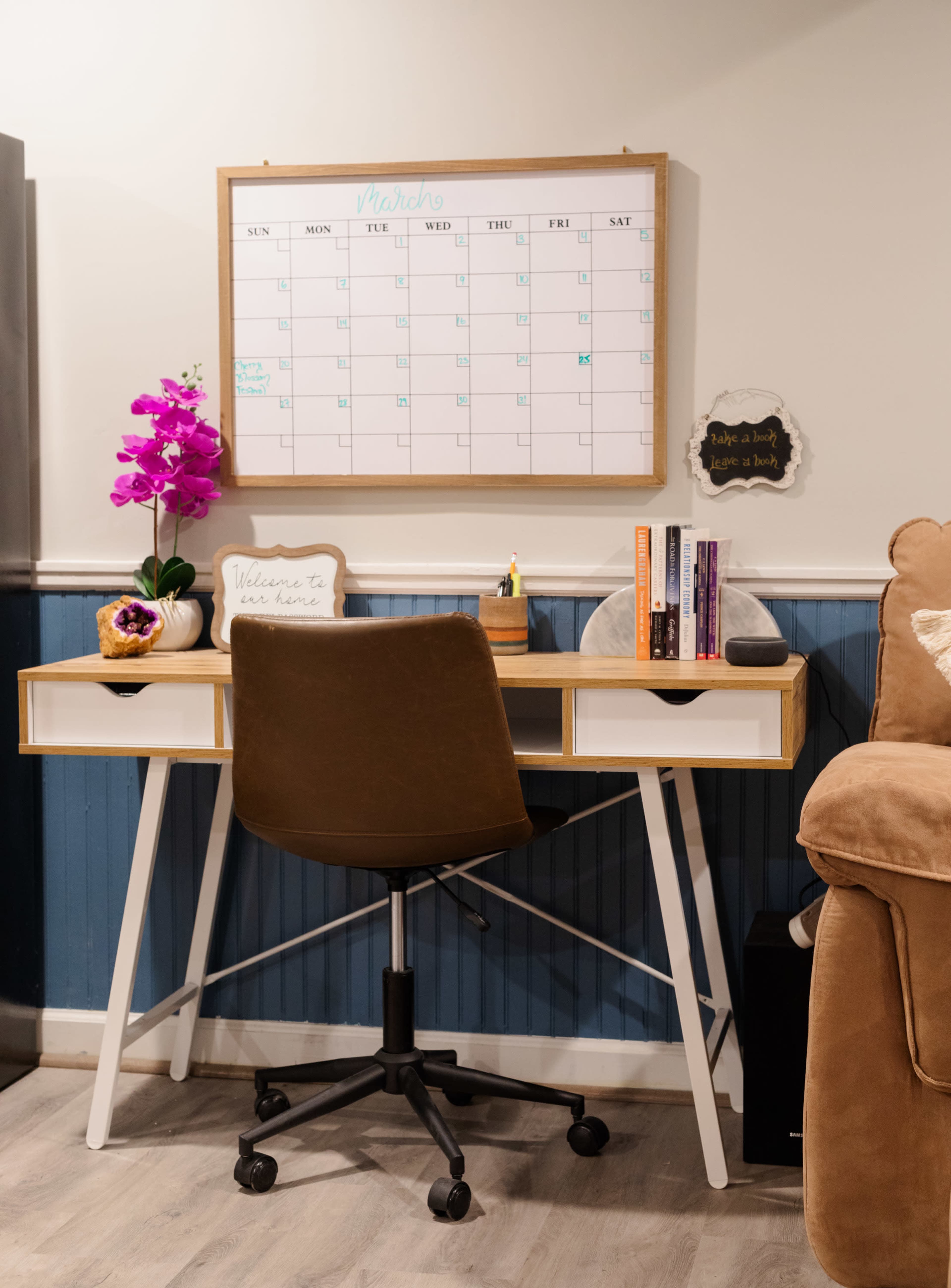 A home office setup features a wooden desk with a swivel chair, a calendar on the wall, and decorative items like books and a plant.