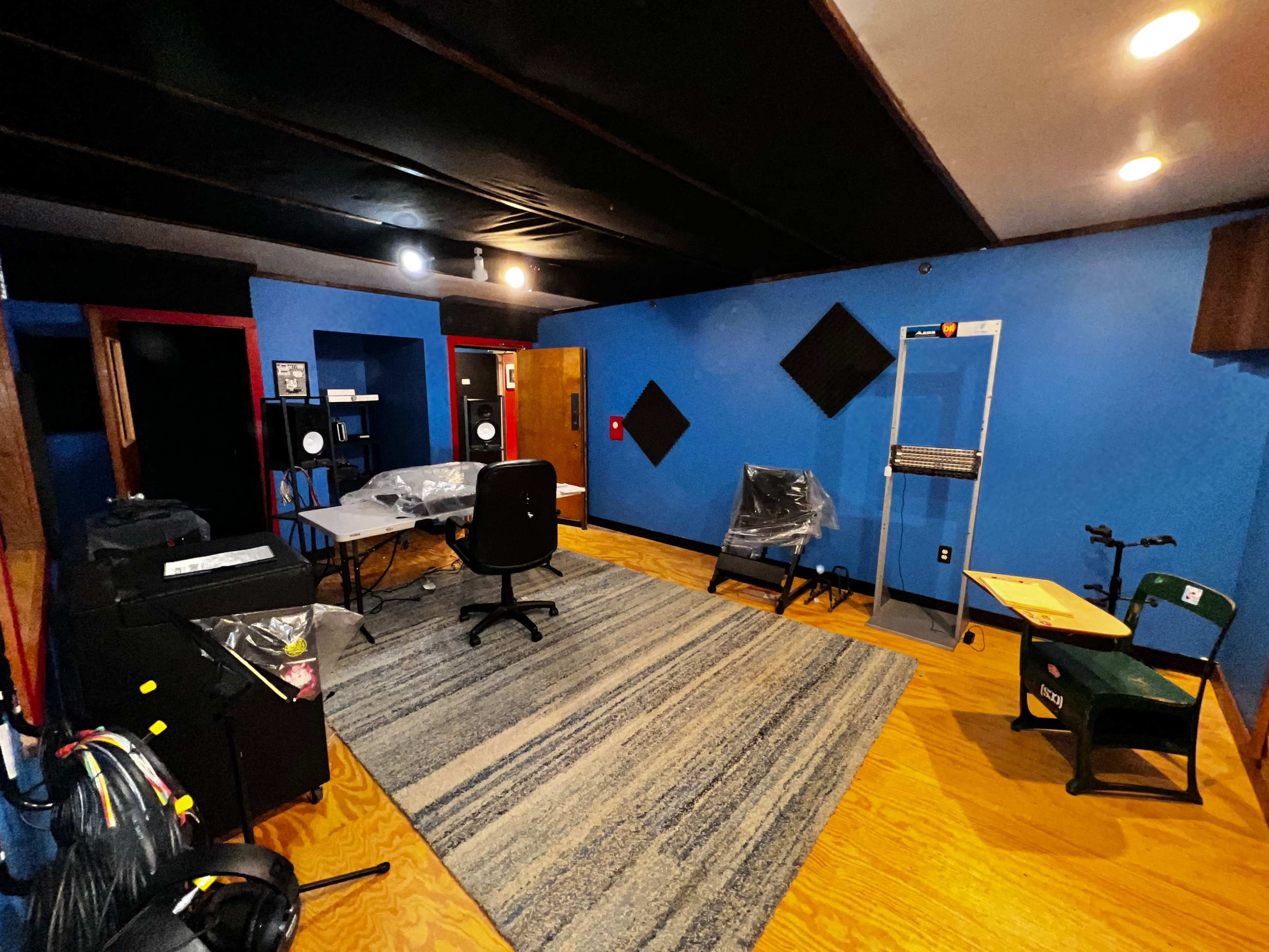 The image shows a spacious room with blue walls, wooden flooring, and various pieces of furniture including a desk, chairs, and soundproofing panels.