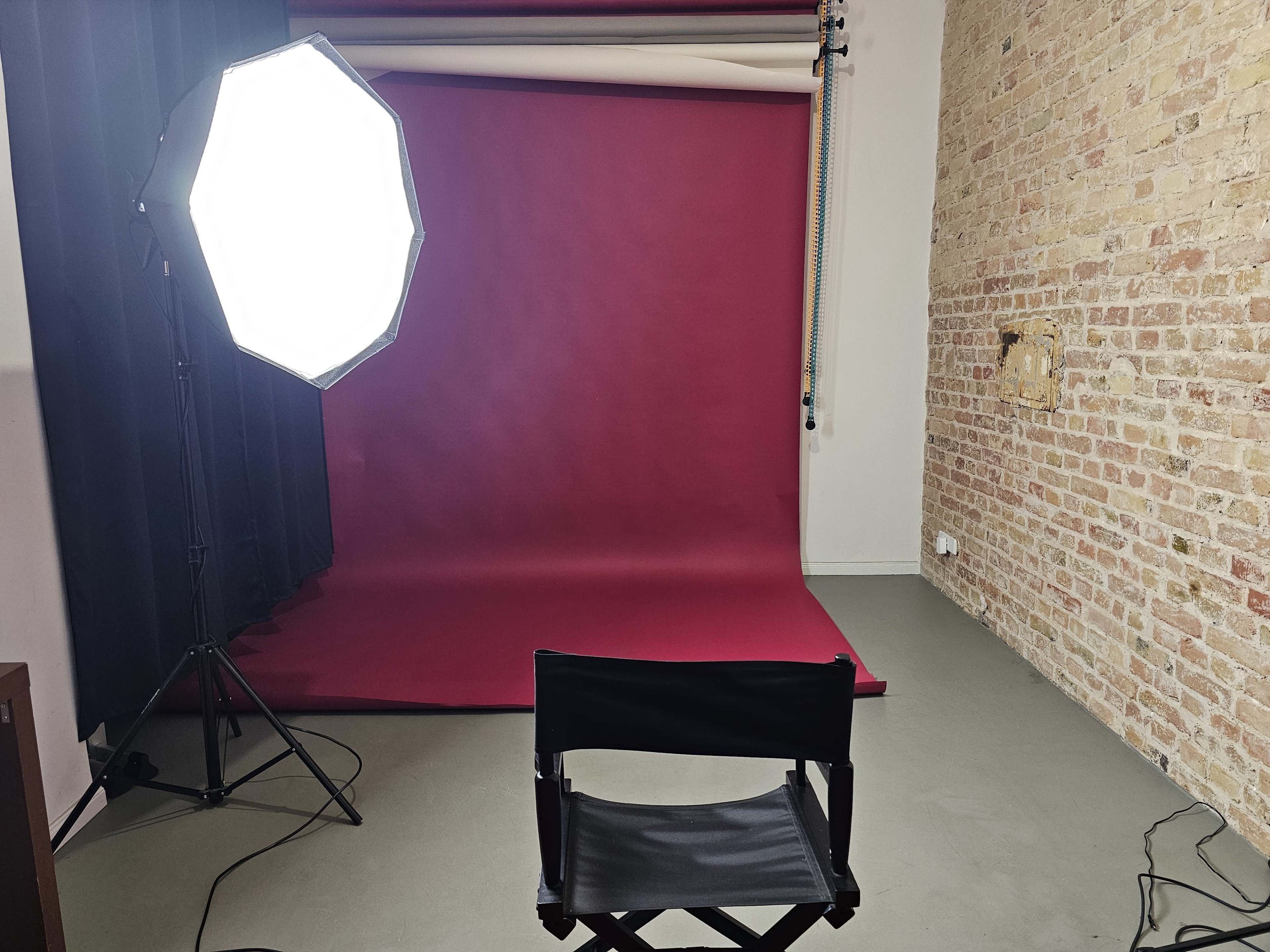 The image shows a photography studio with a maroon backdrop, a large light source, and a director's chair positioned in front of the backdrop.