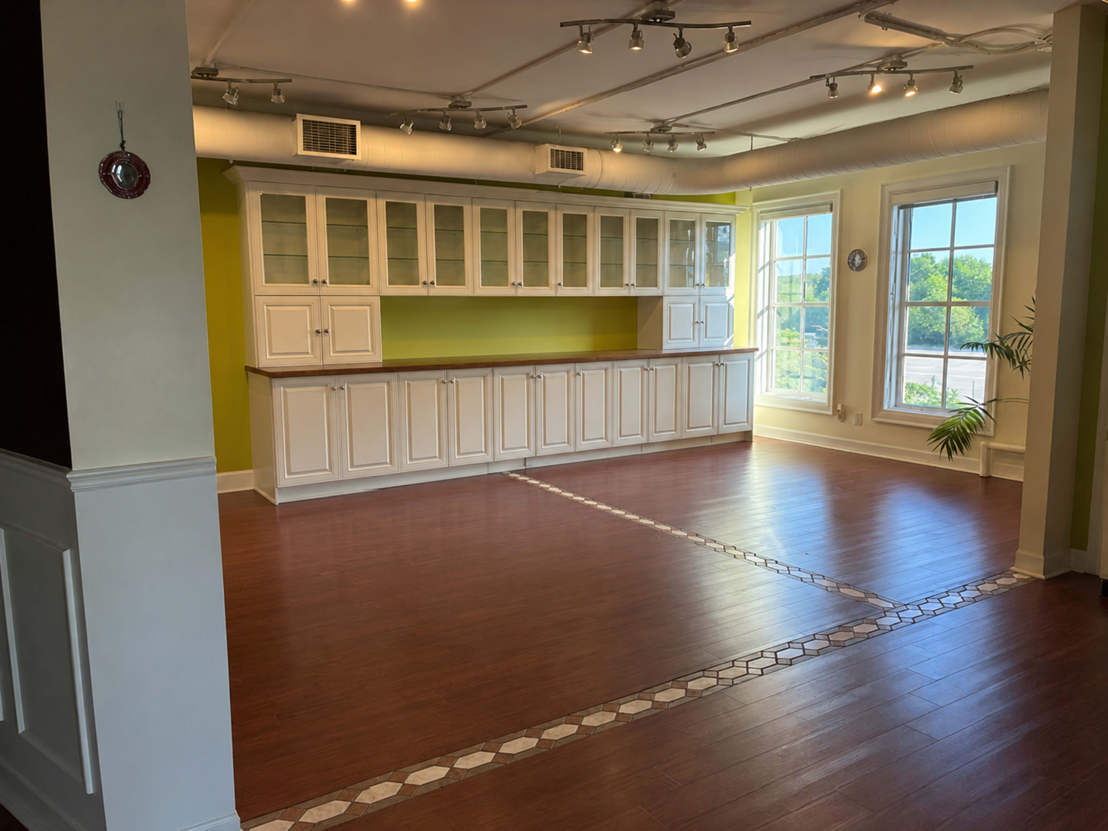 The image shows an empty room featuring a large white cabinetry installation along one wall and a wooden floor with a decorative tile border.