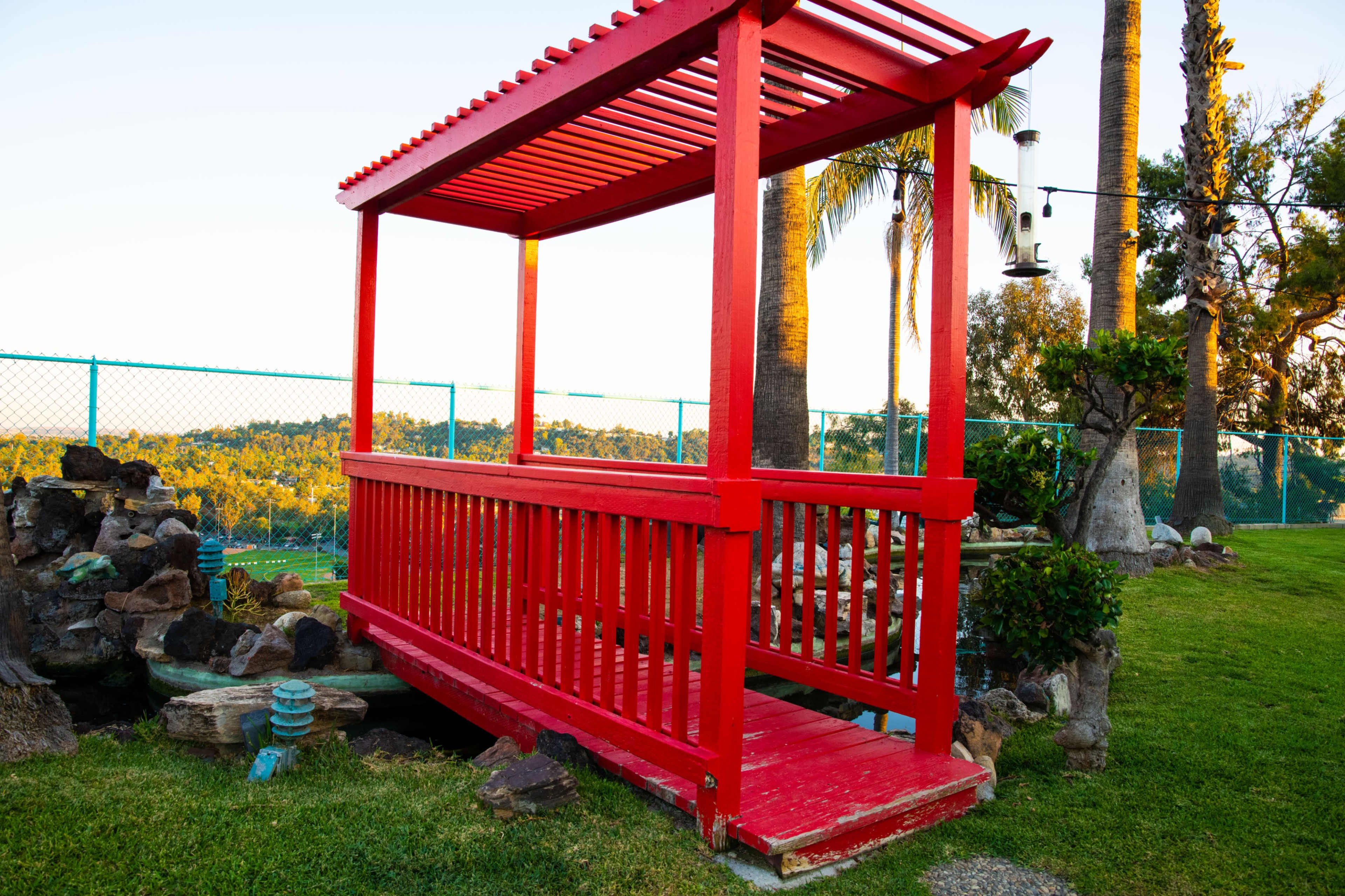 A bright red gazebo with a slatted roof stands on a grassy area surrounded by rocks and palm trees.