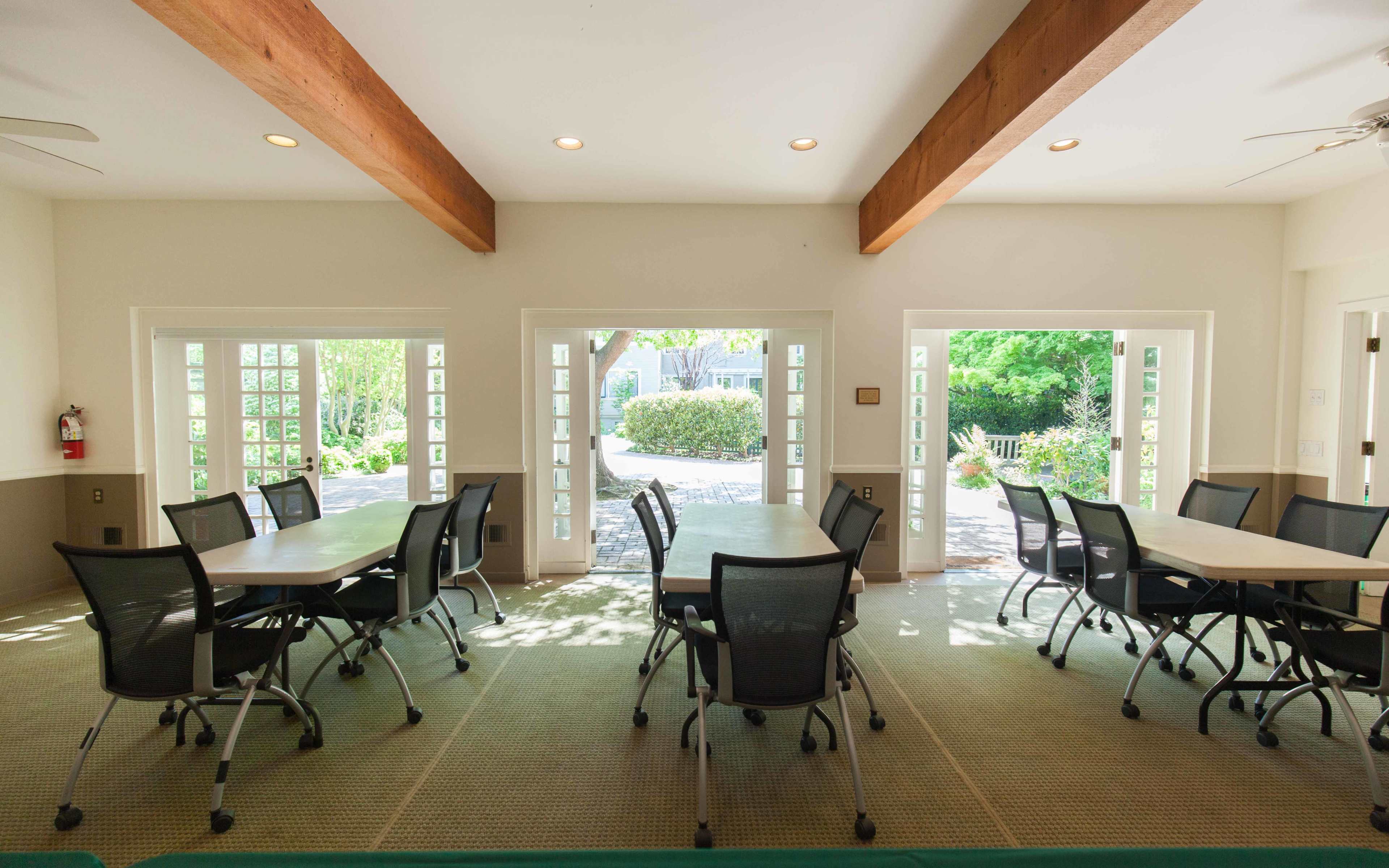 A bright room with wooden beams features multiple round tables surrounded by black mesh chairs, with large windows opening to a garden view.