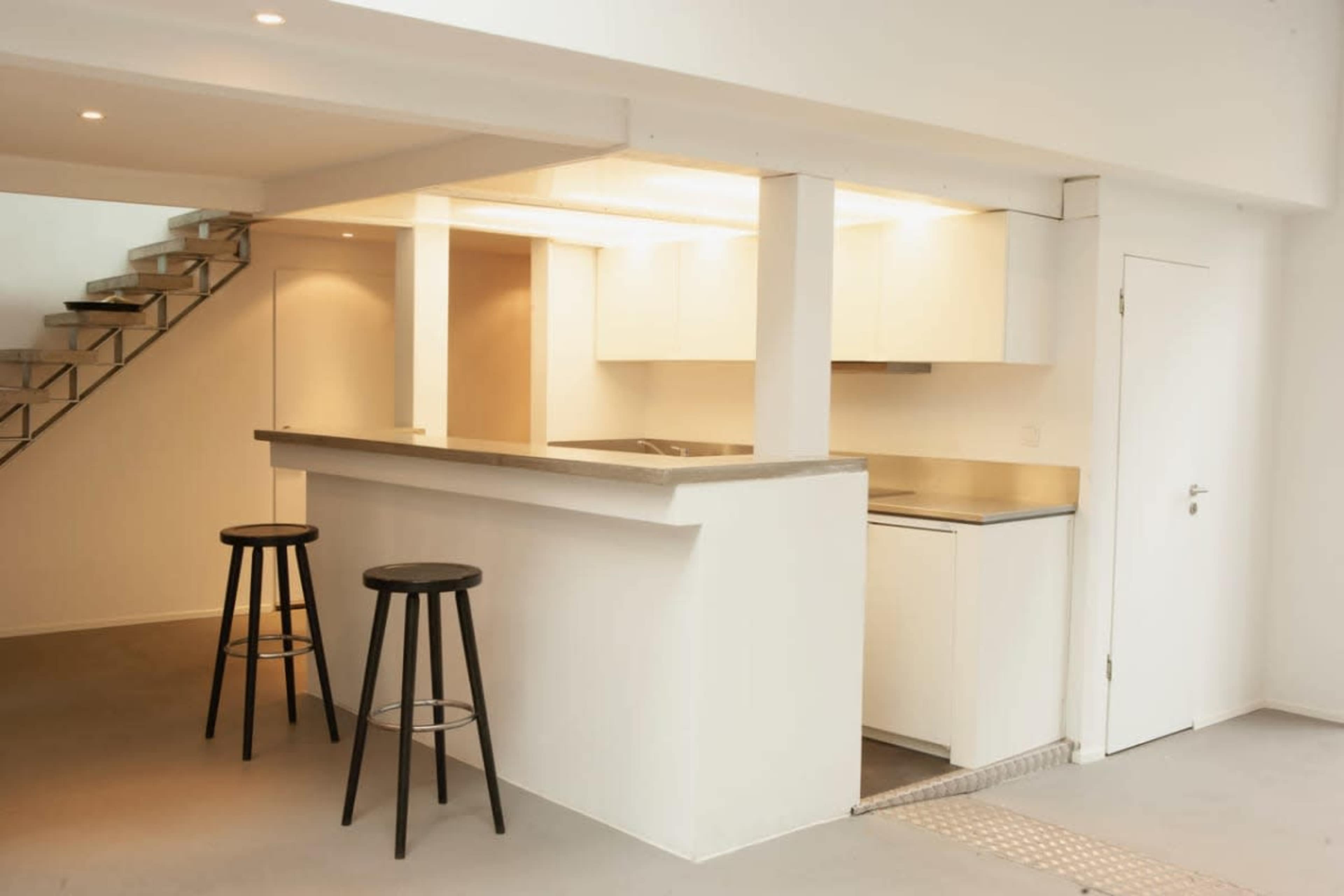 The image shows a modern kitchen area with a bar counter and two black stools, located near a staircase.