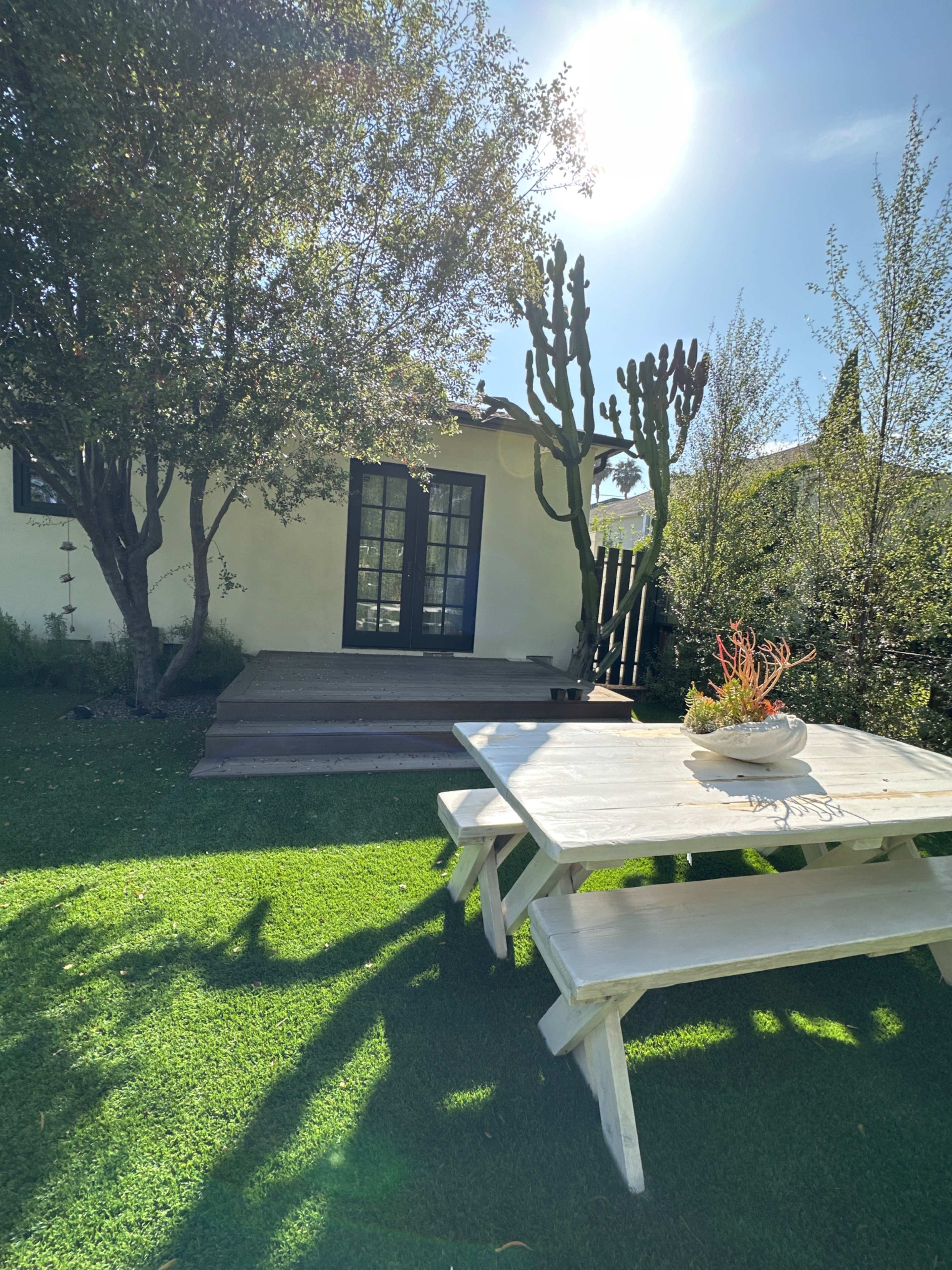 A wooden picnic table sits on a grassy area in front of a small house with a deck and tall cacti nearby.