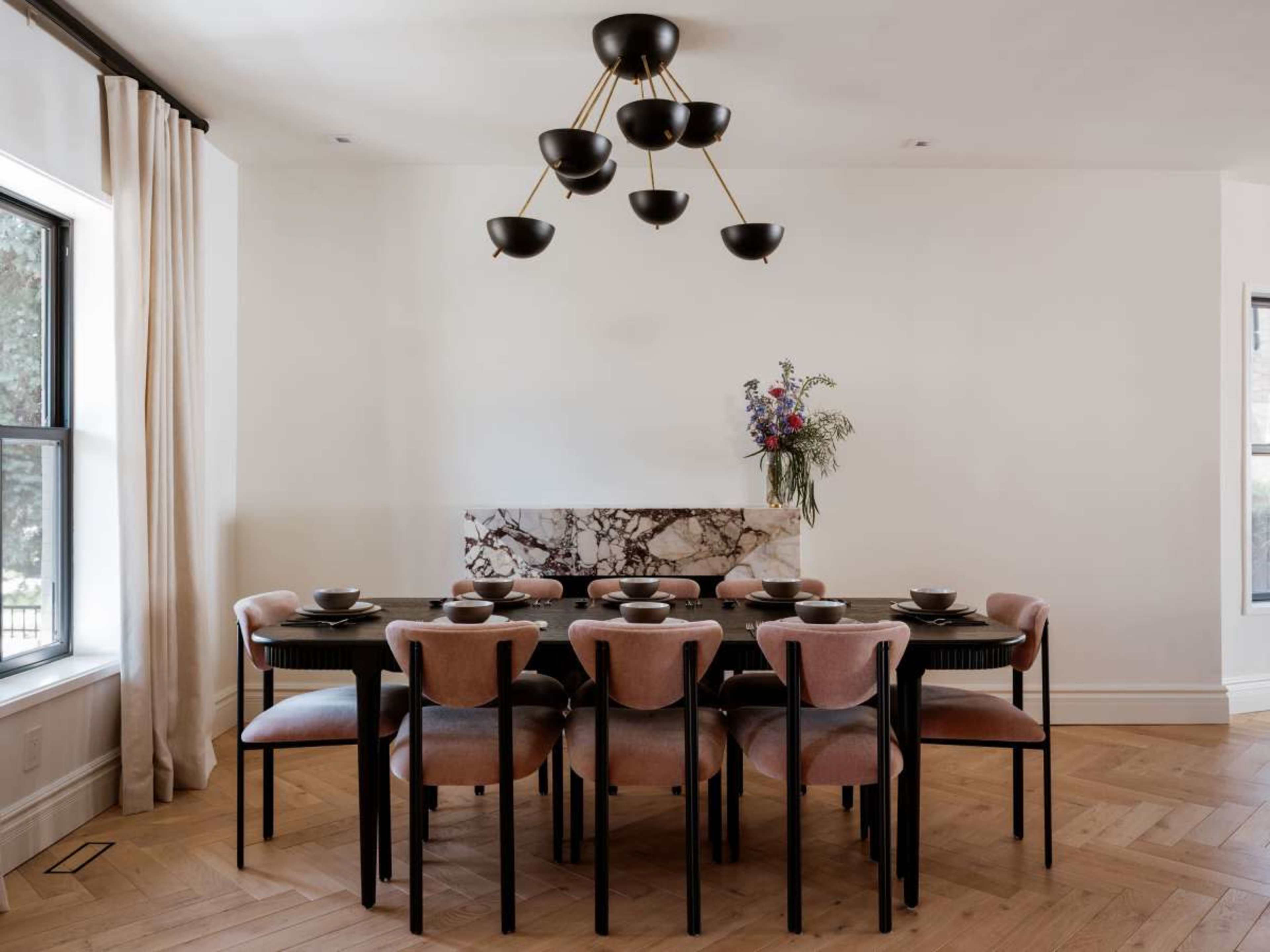 A dining room features a long black table set with dishes, surrounded by upholstered chairs, beneath a modern chandelier and a floral arrangement on the wall.
