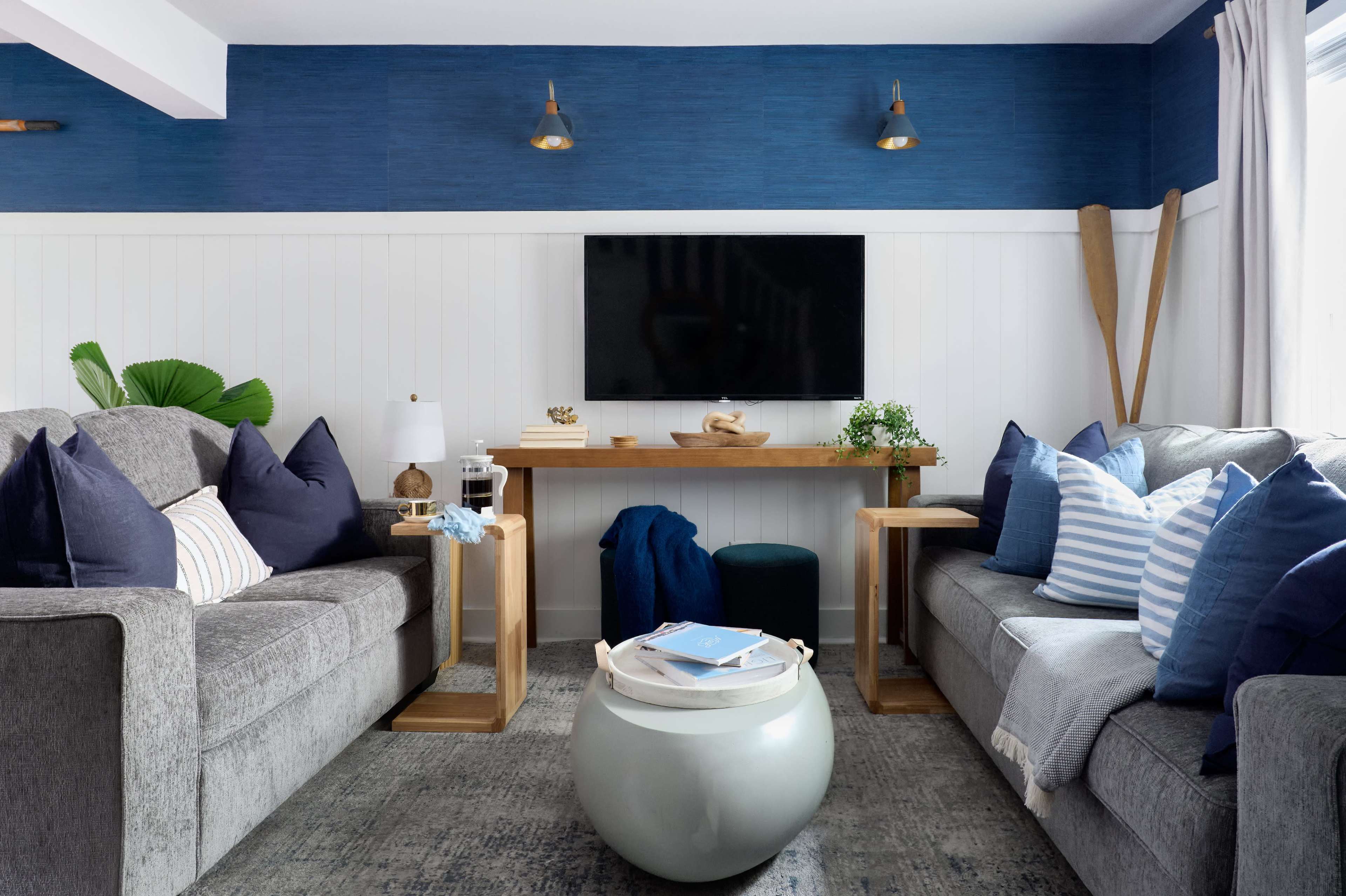 The living room features two gray sofas with numerous blue and white pillows, a wooden console table beneath a wall-mounted television, and decorative elements including a plant and paddles.