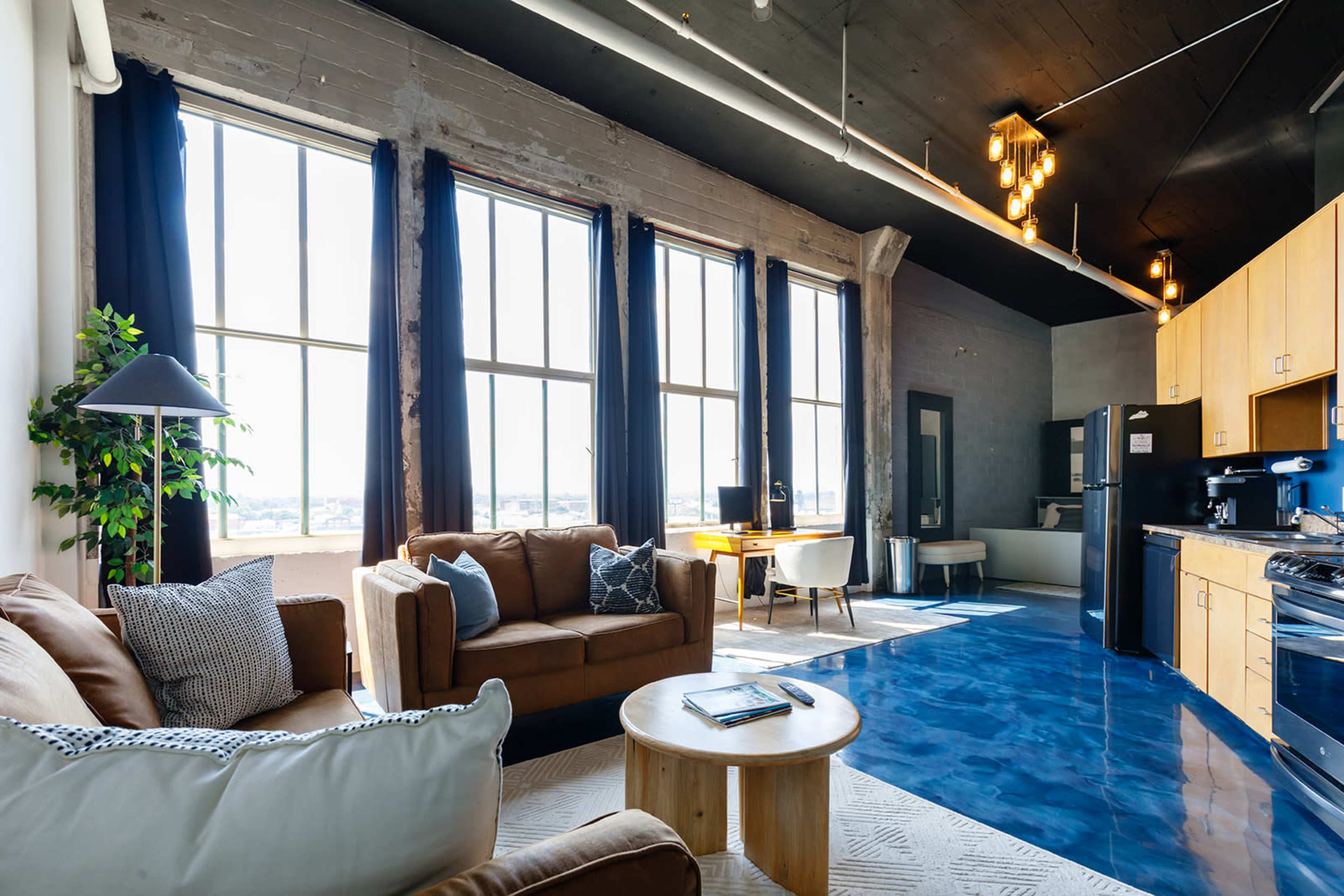 The image shows a modern living space with large windows, a comfortable sofa, a wooden coffee table, and a kitchen area in the background.