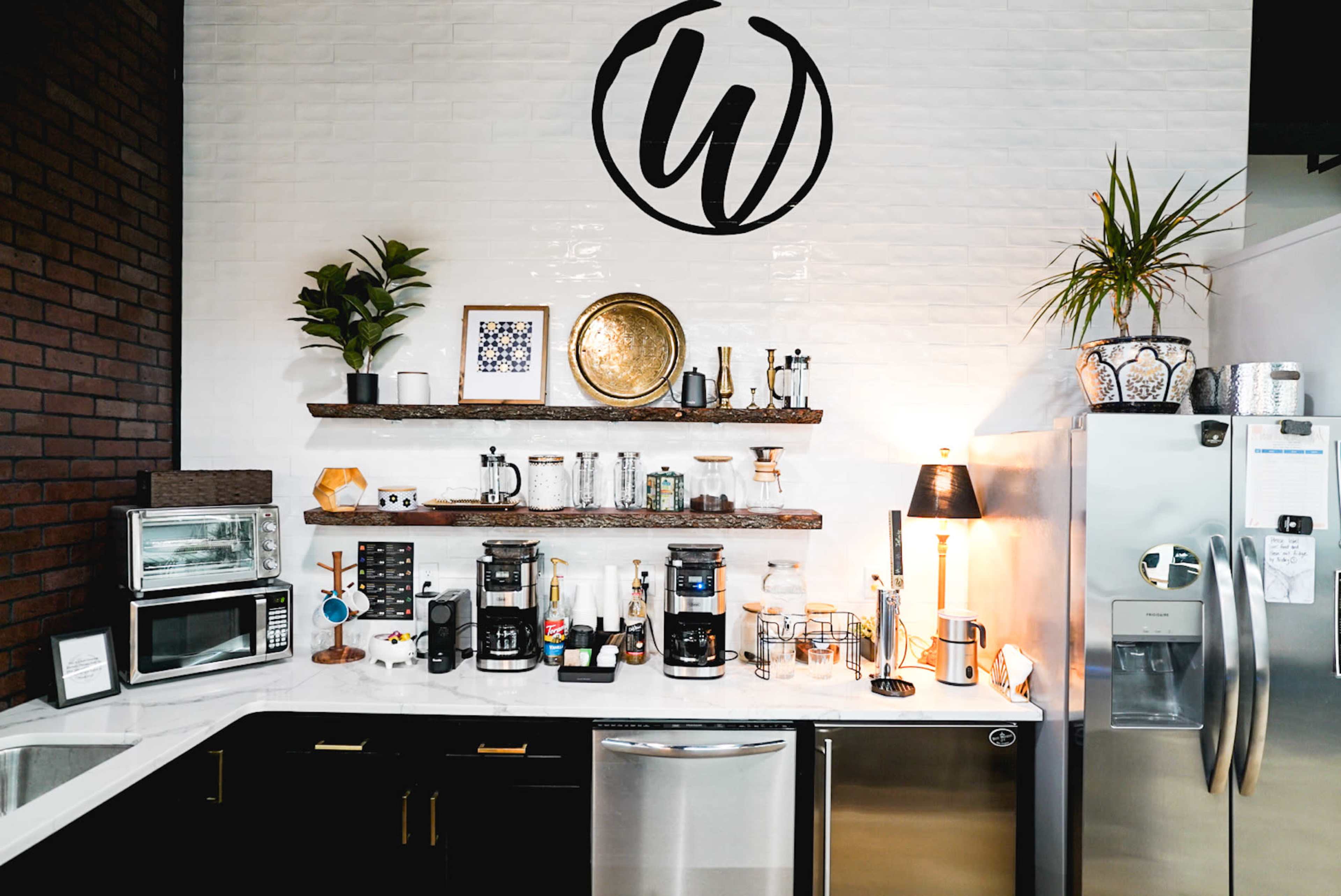The image depicts a modern kitchen setup with a collection of appliances and decorative items arranged on a countertop and shelves against a white and brick wall.