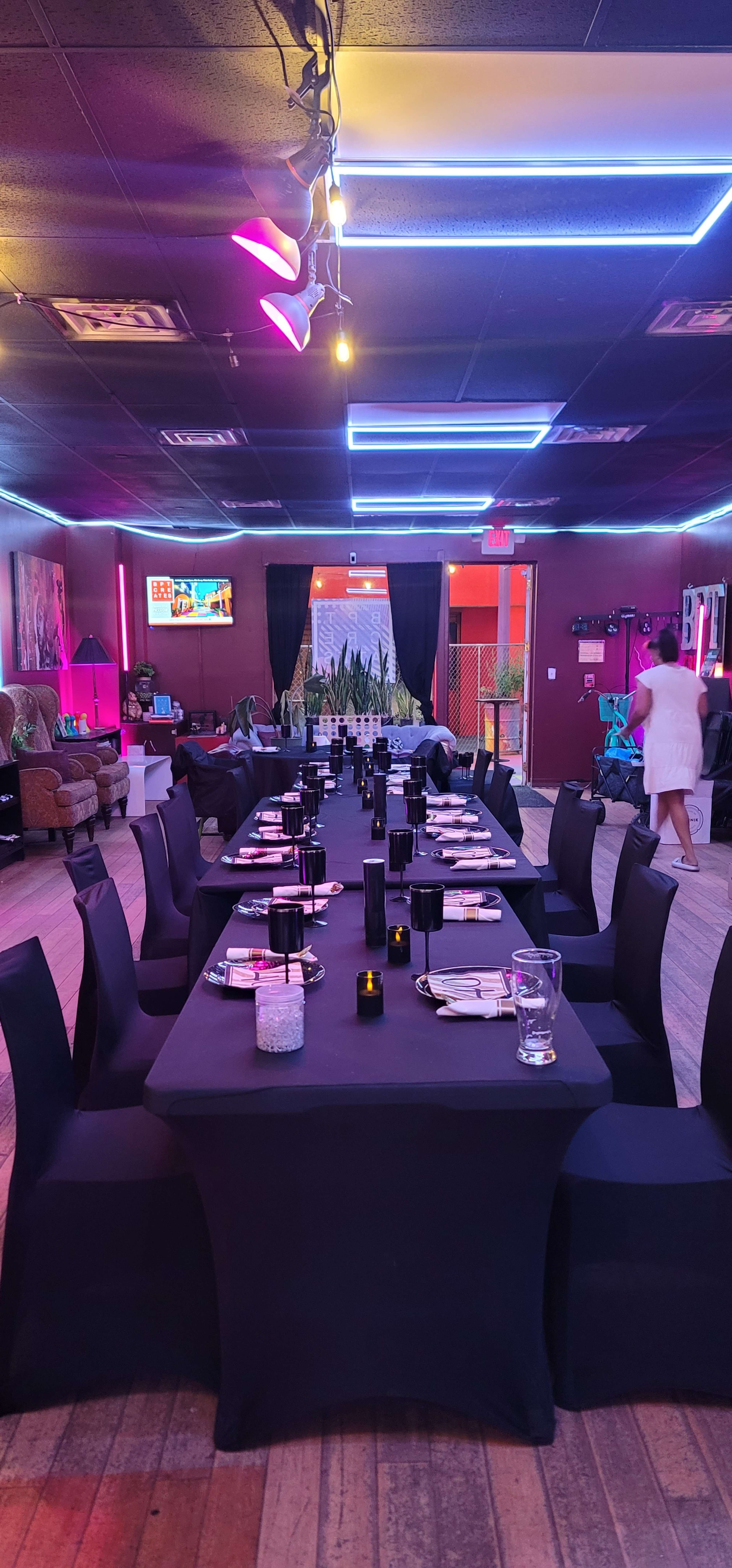 A long, elegantly set dining table with black tablecloths and dark decor is positioned in a brightly lit restaurant featuring neon lighting and vibrant colors.