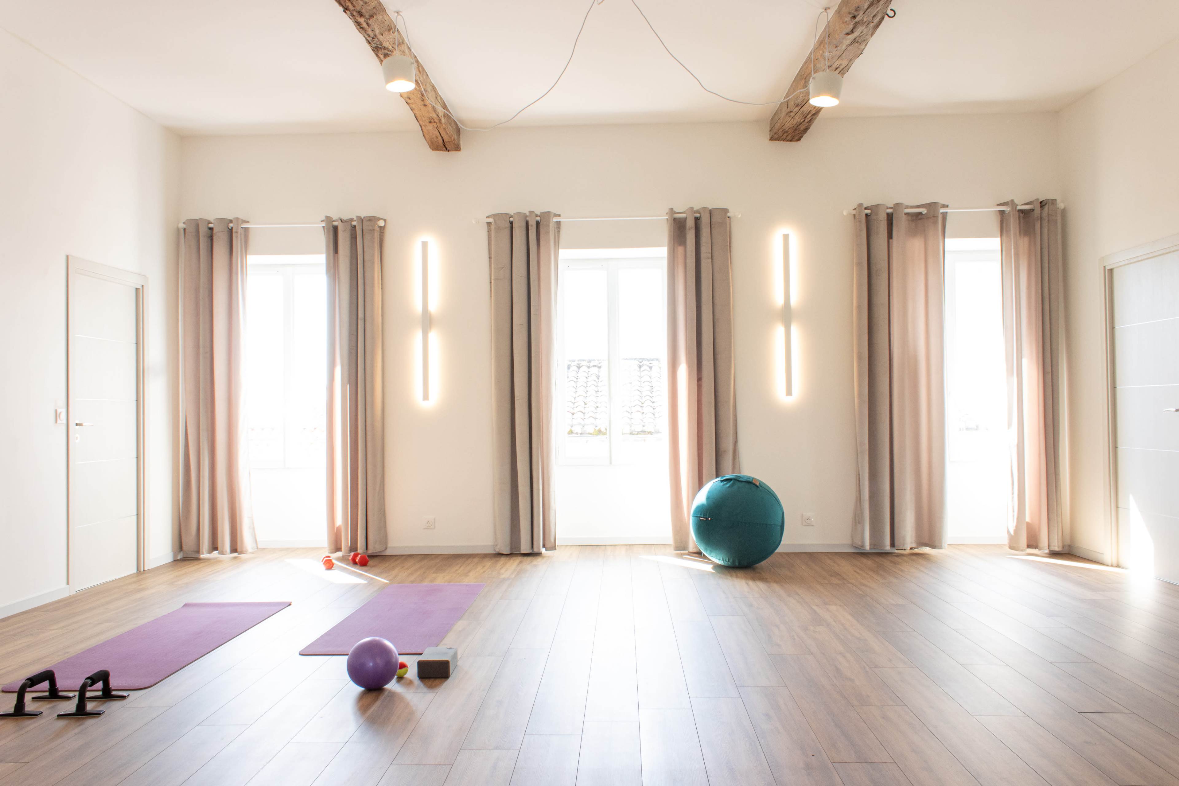 The room features wooden flooring, large windows with curtains, and exercise equipment including mats, a stability ball, and weights.