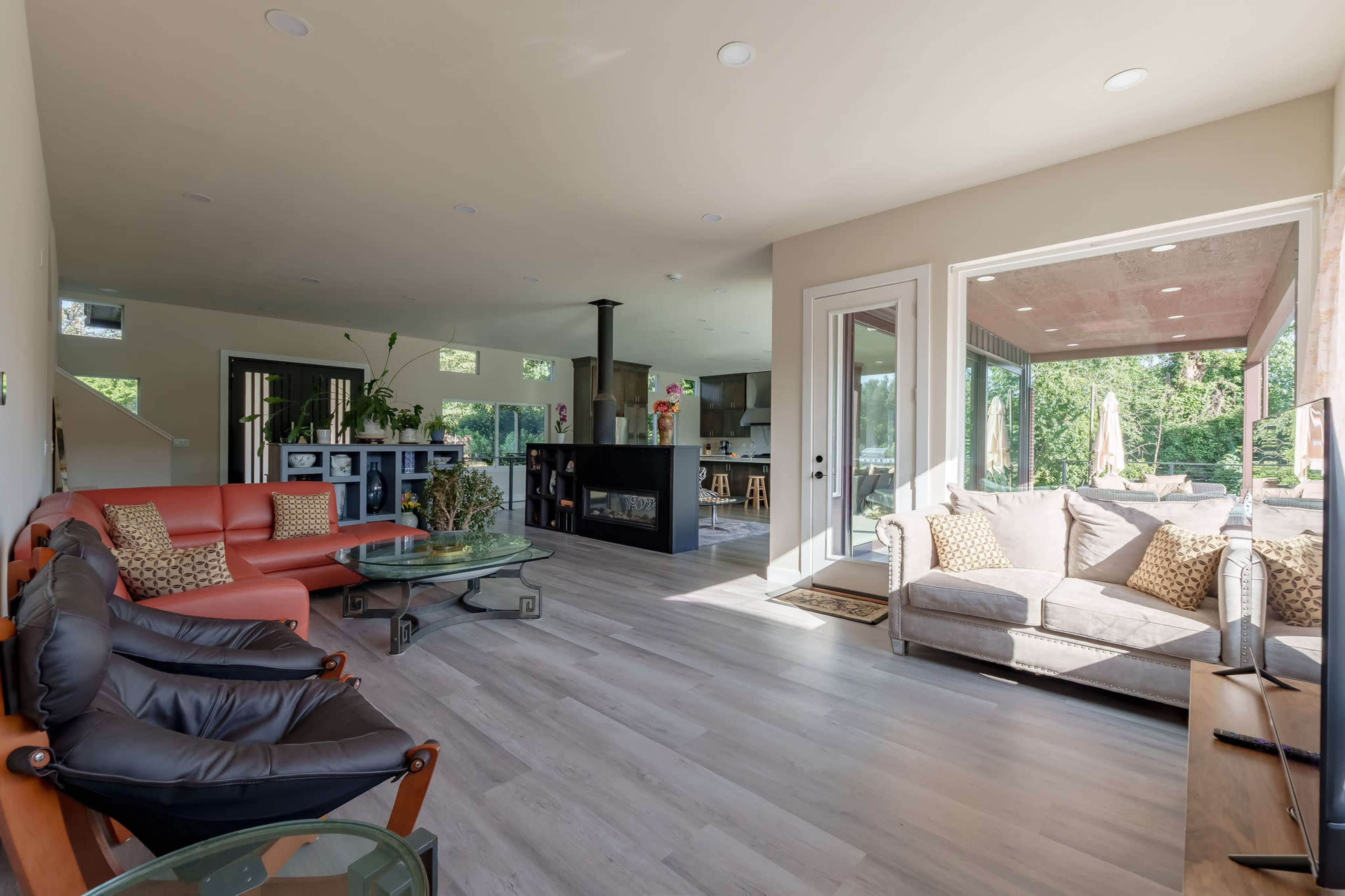 The image shows a modern living room with a sectional sofa, armchairs, a glass coffee table, and large windows that open to a patio area.