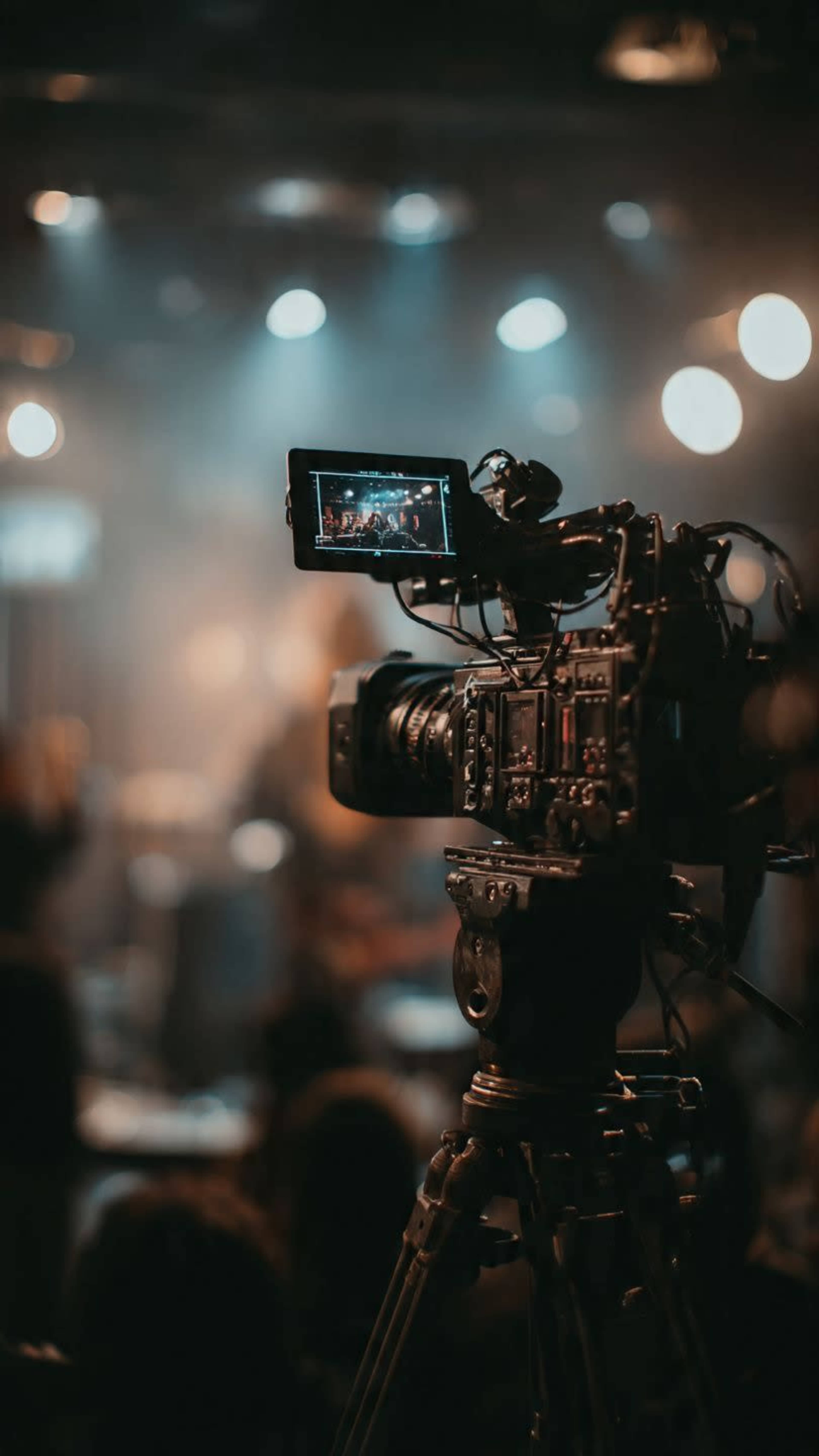 A video camera on a tripod captures a performance in a dimly lit venue with blurred silhouettes in the background.