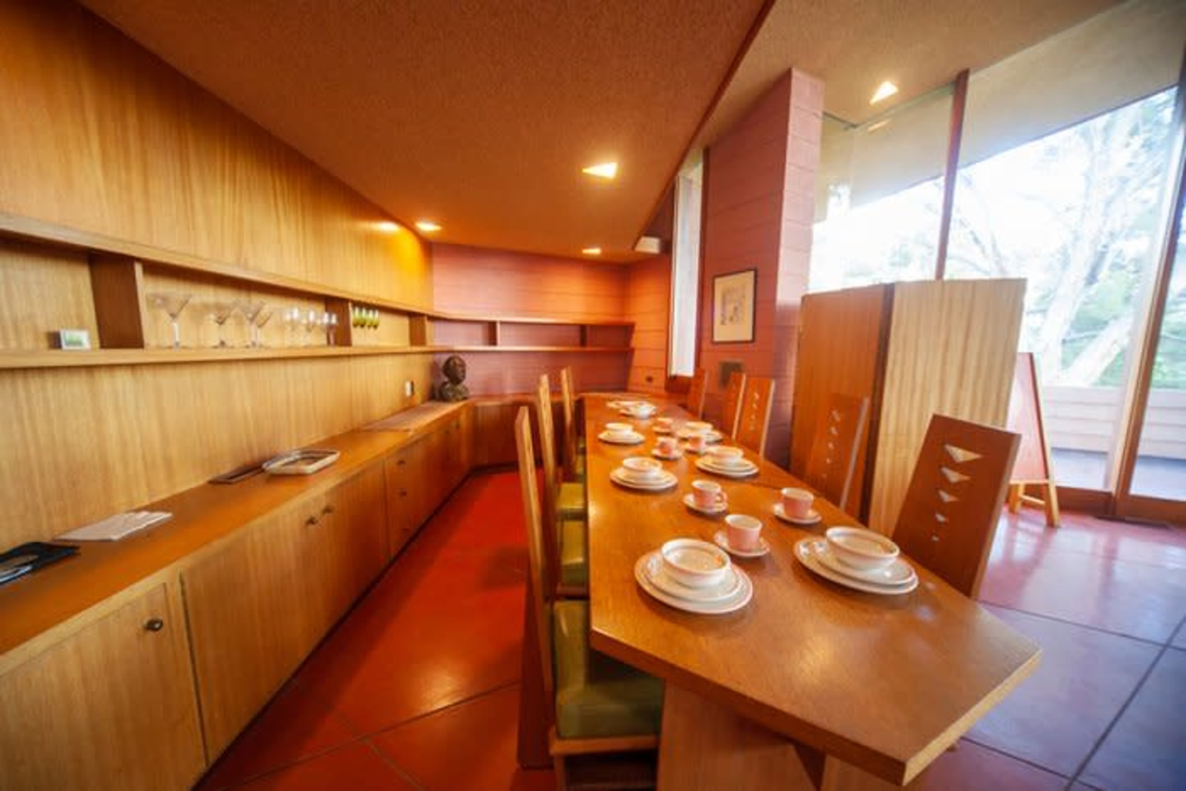 A modern dining area features a wooden table set with plates and cups, surrounded by green chairs against a backdrop of wooden cabinetry and large windows.