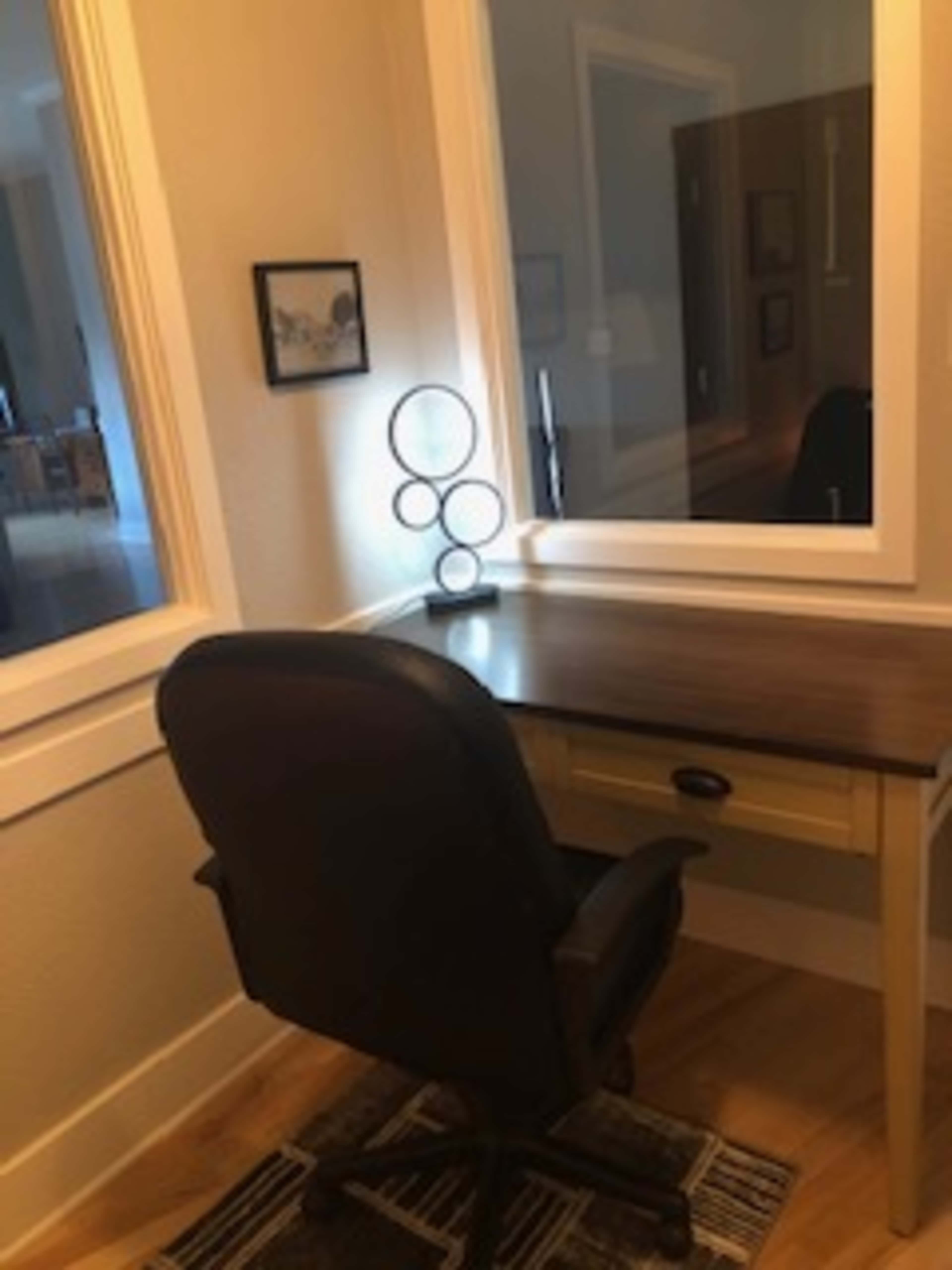 A simple desk and an office chair positioned beside a mirror, with a circular lamp on the surface.